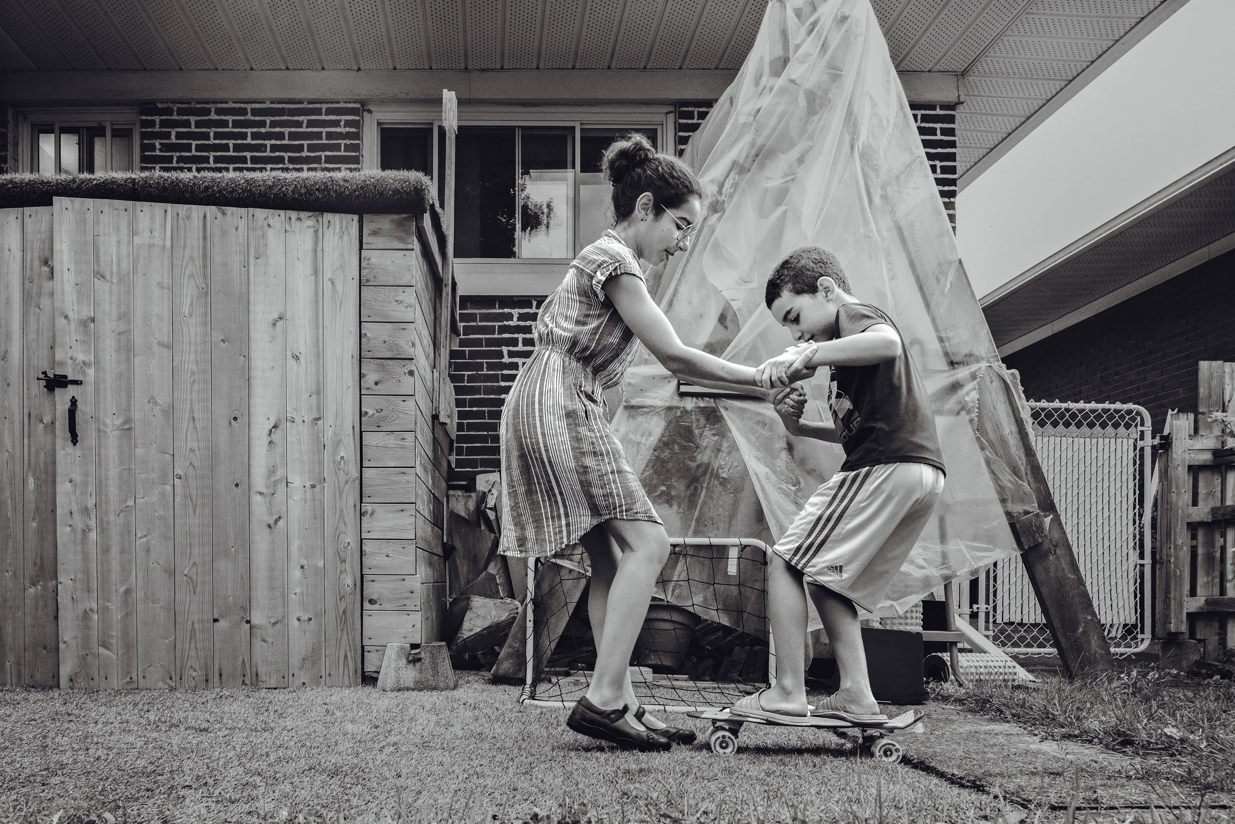 A woman and boy are outside, balancing on a skateboard. The woman is assisting the boy as he tries to stand on the skateboard, both holding onto each other's hands. There is a large plastic sheet draped over a structure behind them, and a wooden fenc