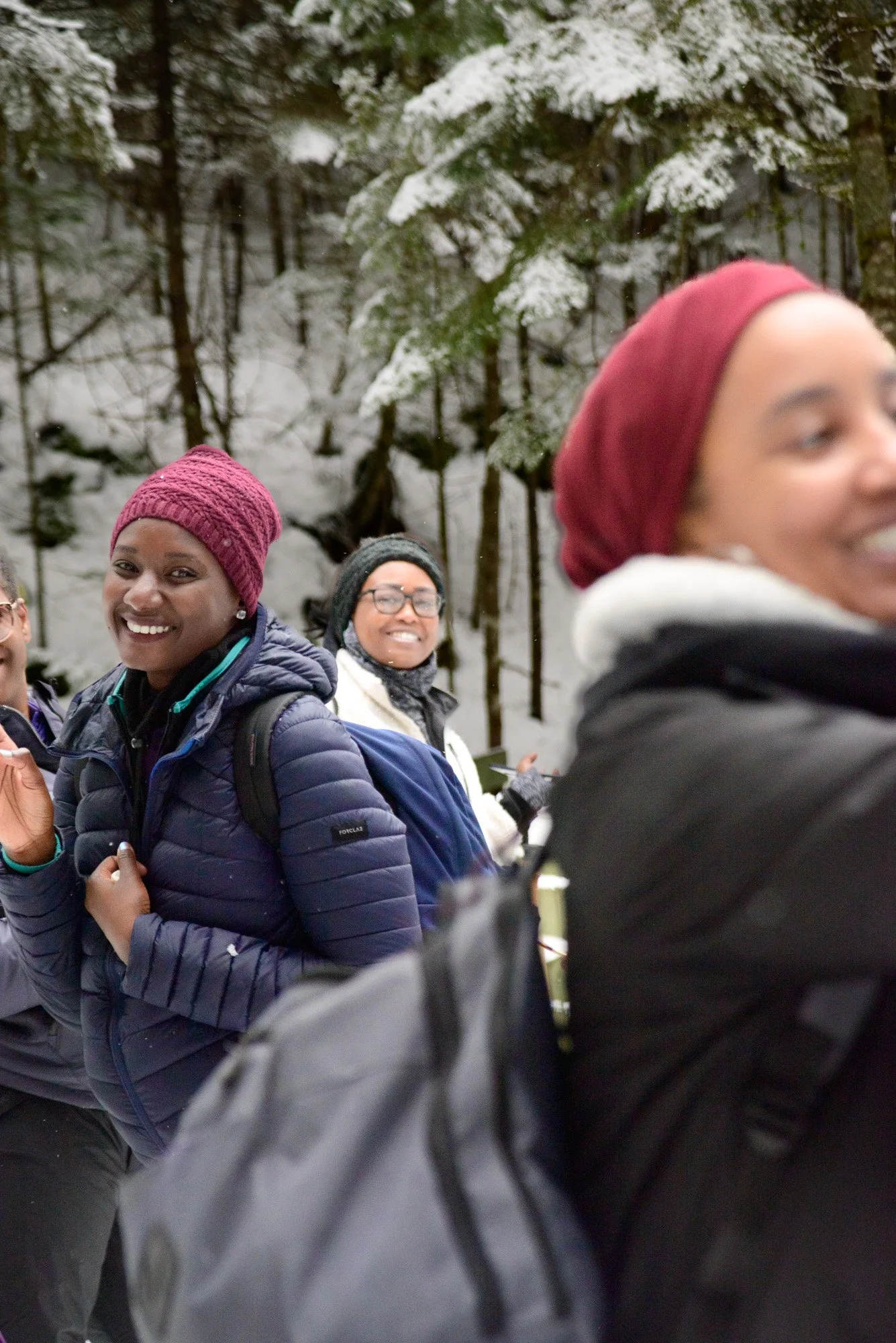 Group of four women smiling outdoors in a snowy forest, wearing winter coats and hats.