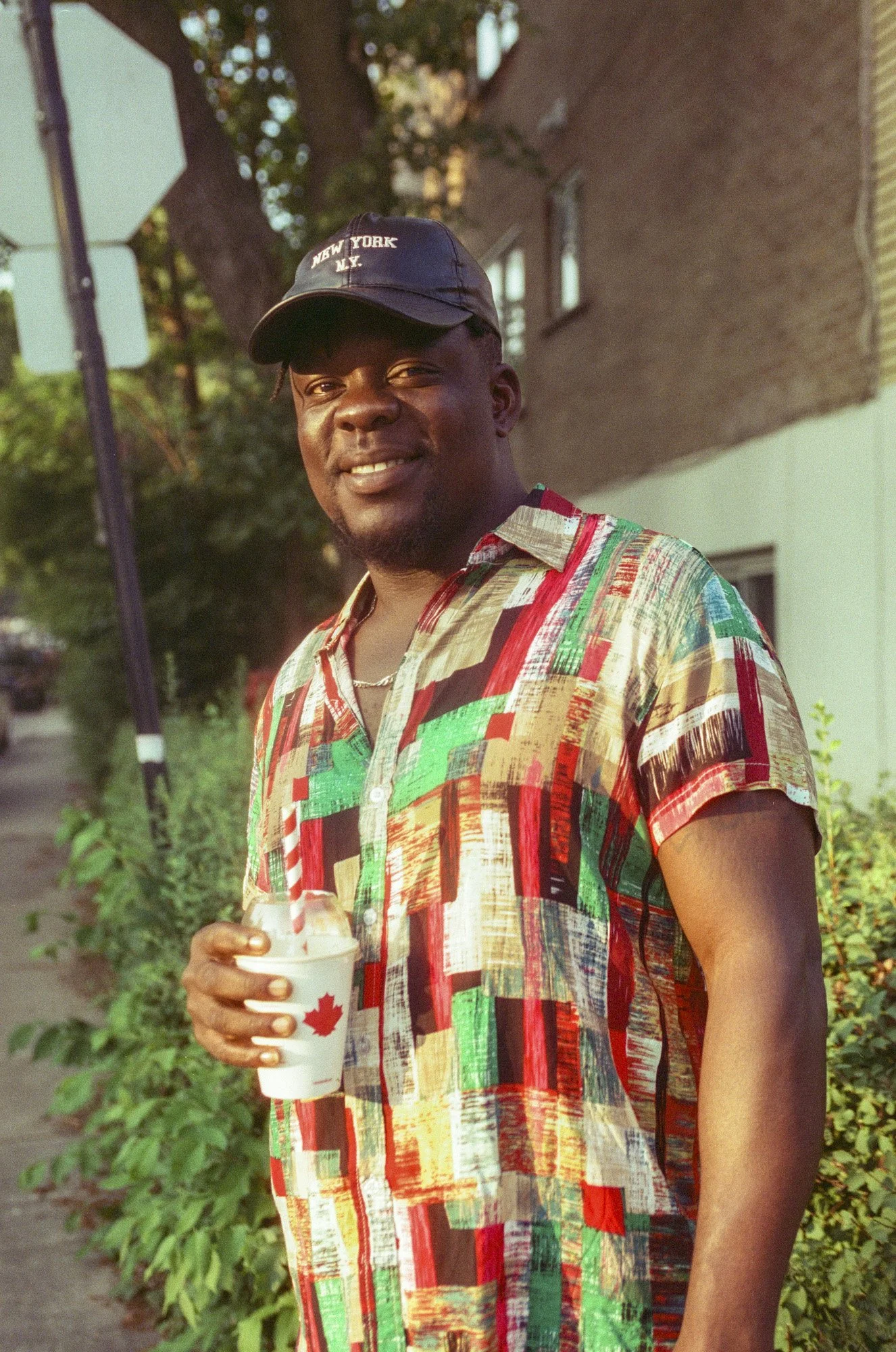 A smiling man wearing a black New York, NY cap and a colorful patchwork shirt, holding a white cup with a red maple leaf and a straw, standing outdoors near greenery and a house during daylight.