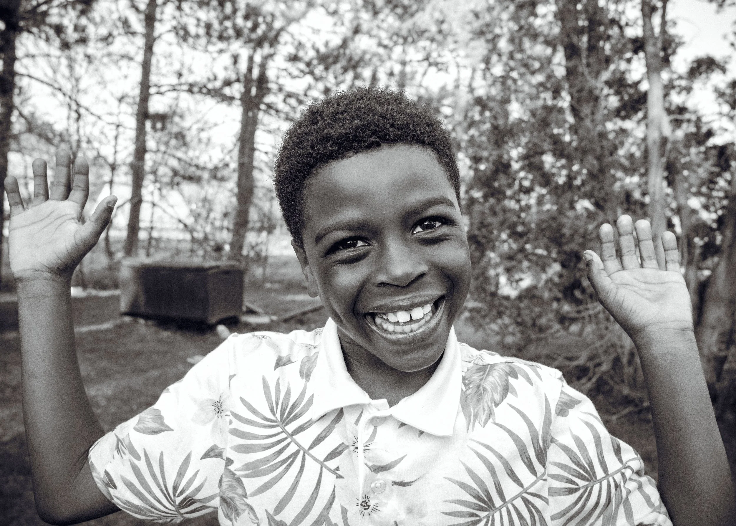 A smiling woman with short curly hair, wearing a patterned shirt, raising her hands in a playful gesture outdoors with trees in the background.