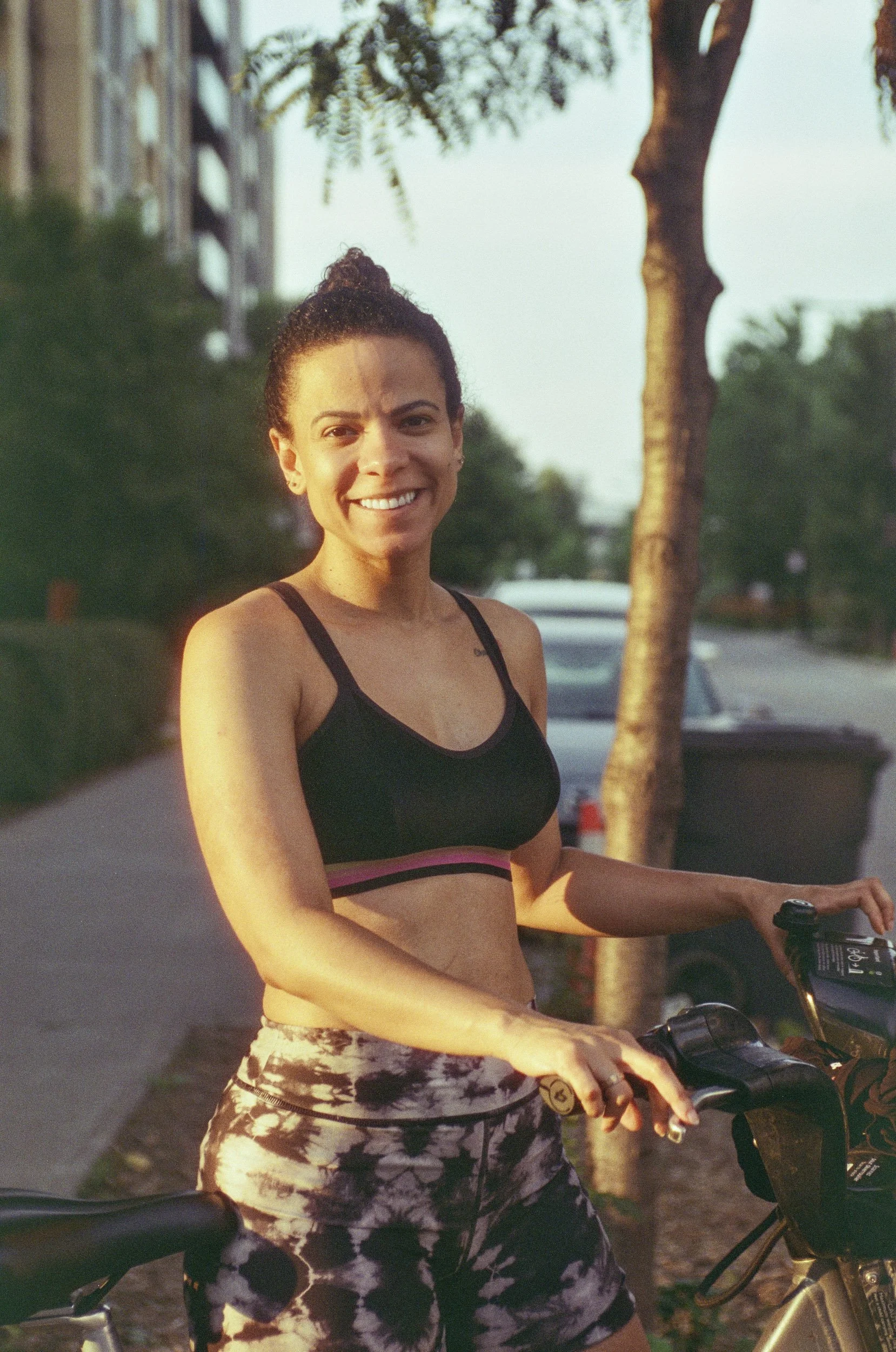 A woman smiling and standing outdoors next to her bicycle during what appears to be late afternoon or early evening. She is wearing a black sports bra and patterned leggings, with her hair tied in a bun. She is holding the handlebar of the bike with 