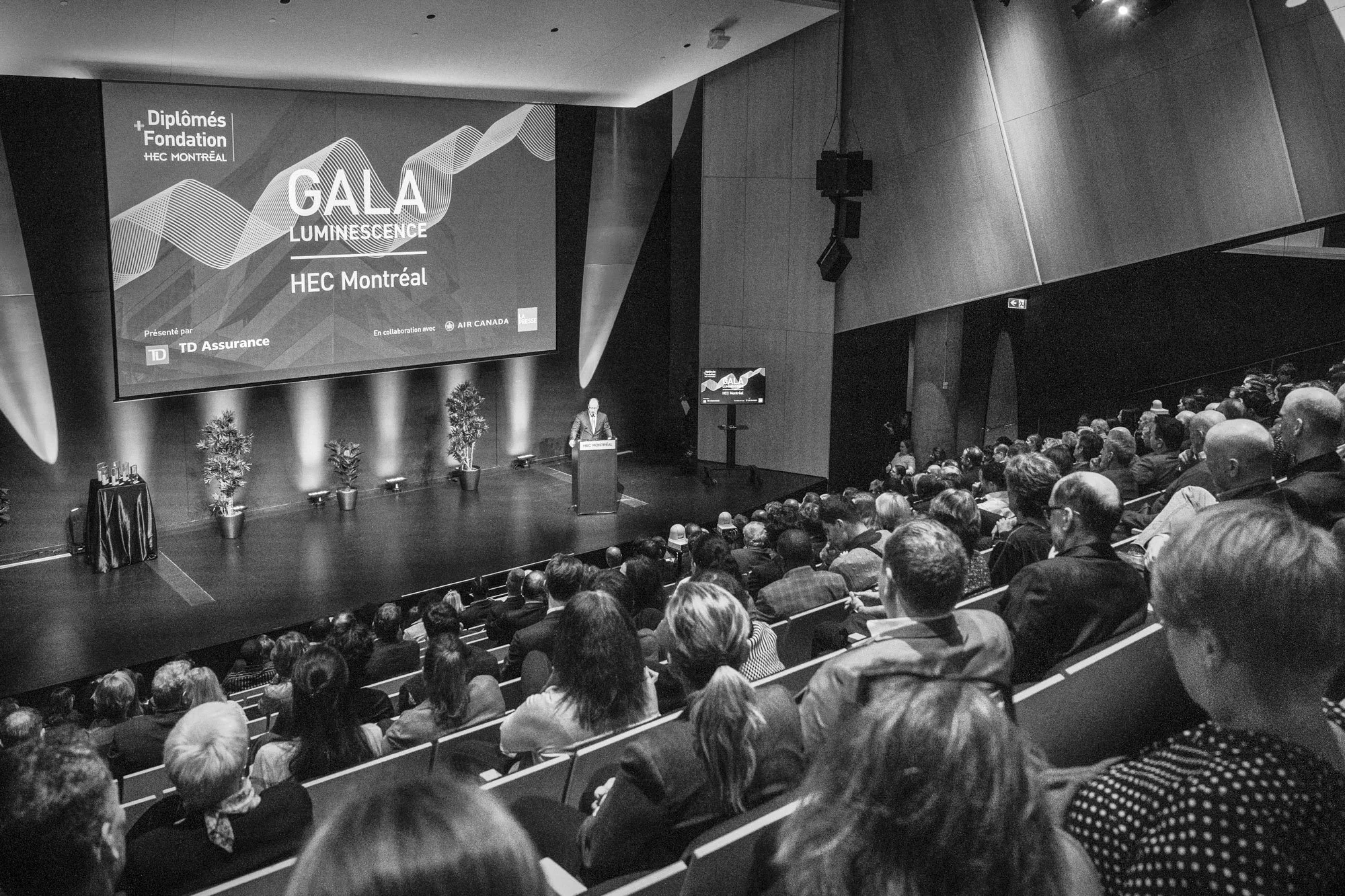A large audience is seated in a modern auditorium during a formal event or conference. A speaker is standing at a podium on stage, giving a presentation. Behind the speaker, a large screen displays the event title 'GALA LUMINESCENCE' along with spons