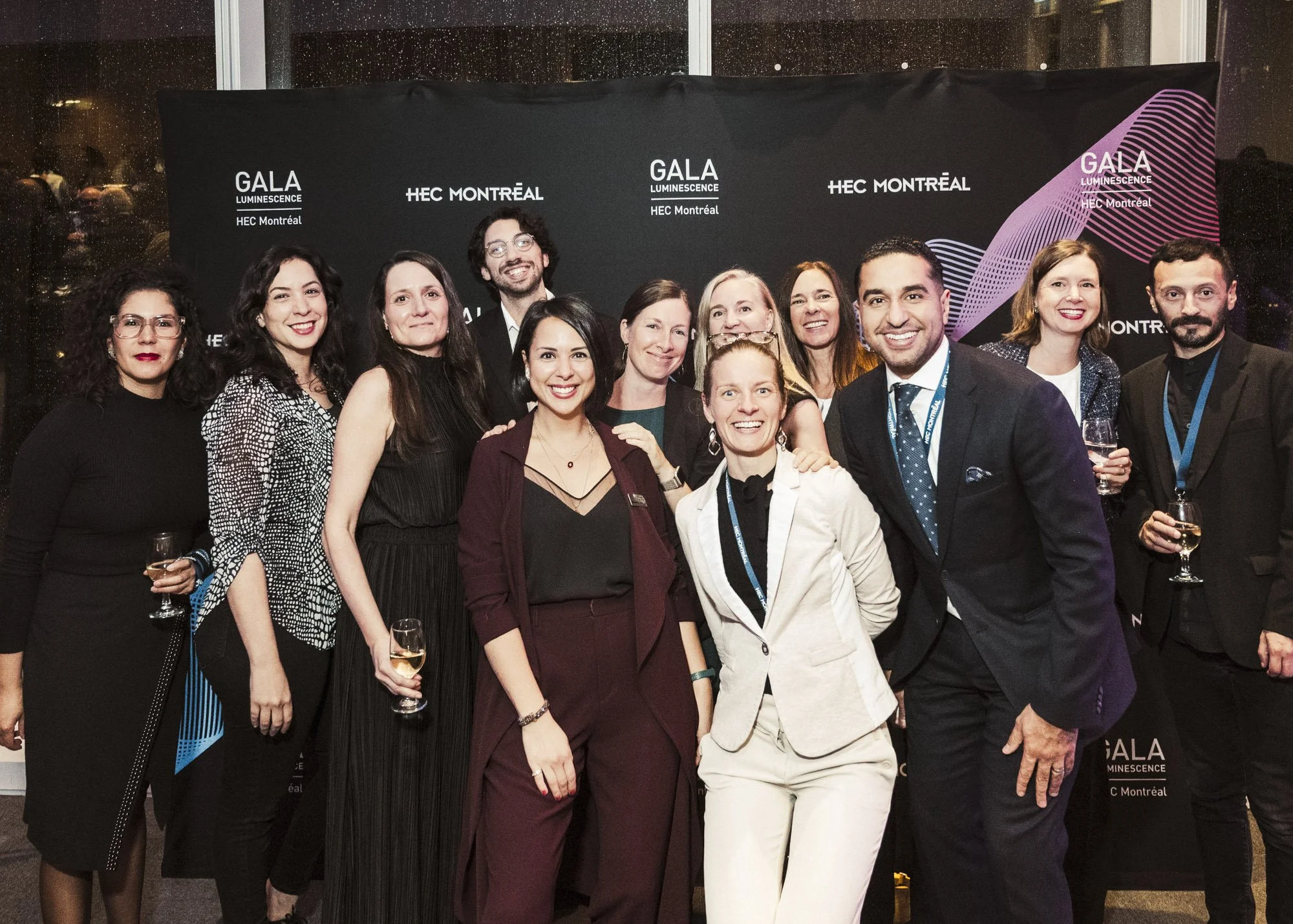 Group of diverse professionals dressed in formal attire, standing together at an event with a black backdrop featuring the words "GALA LUMINESCENCE HEC Montréal." Some are holding glasses of wine and smiling at the camera.