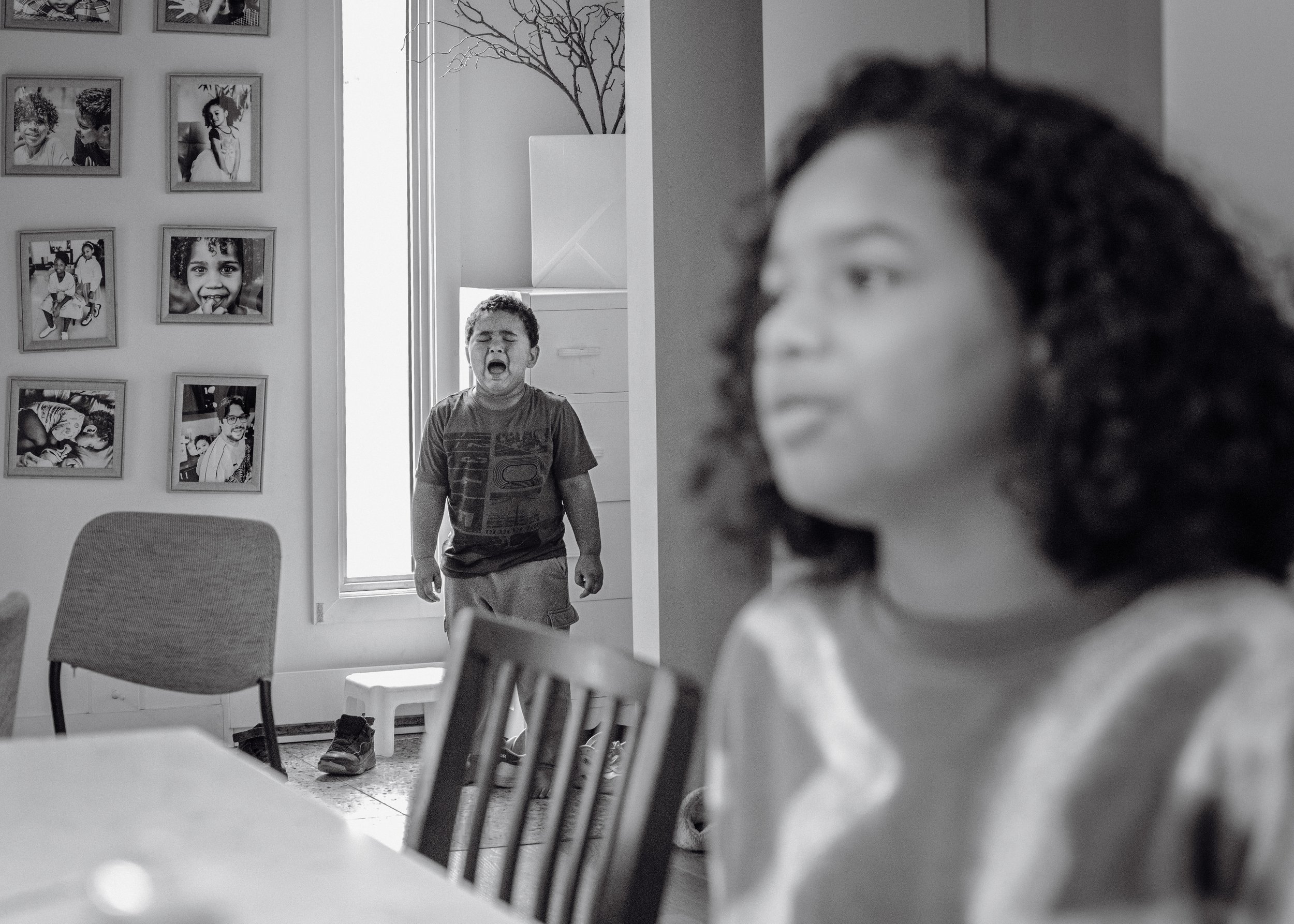 Two children, a boy and a girl, are inside a home. The boy is standing near a door with a window, crying with his eyes closed and mouth open. The girl, with curly hair, is sitting at a table in the foreground, looking to the side. The wall behind the