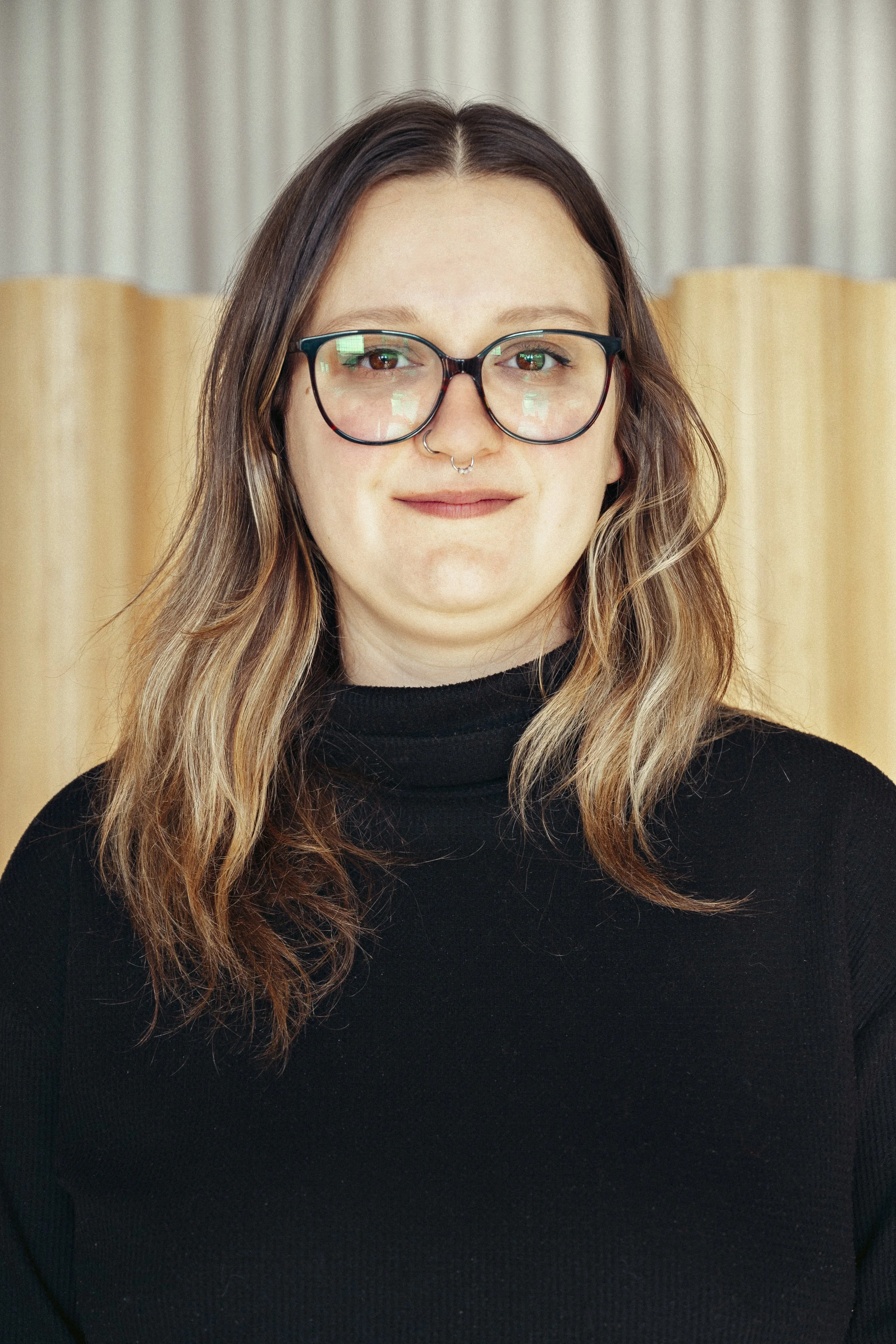 A young woman with long wavy hair, glasses, and a septum piercing, smiling and wearing a black turtleneck in front of a beige curtain.