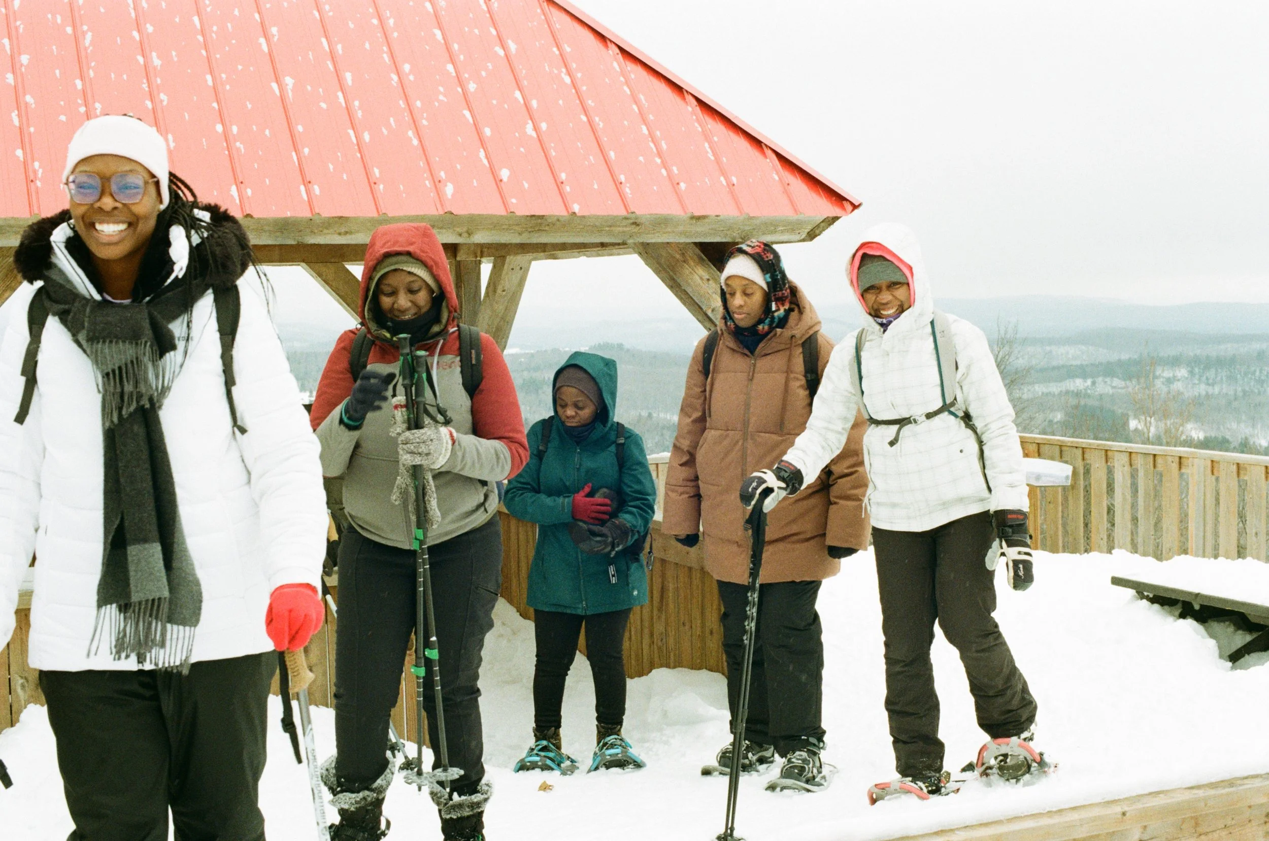 Group of people in winter clothing, standing with snowshoes and trekking poles, in front of a red wooden shelter on a snowy landscape.