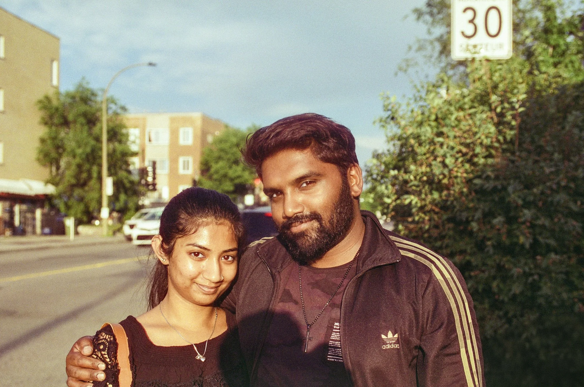 A man and woman posing together outdoors on a street with trees and buildings in the background.