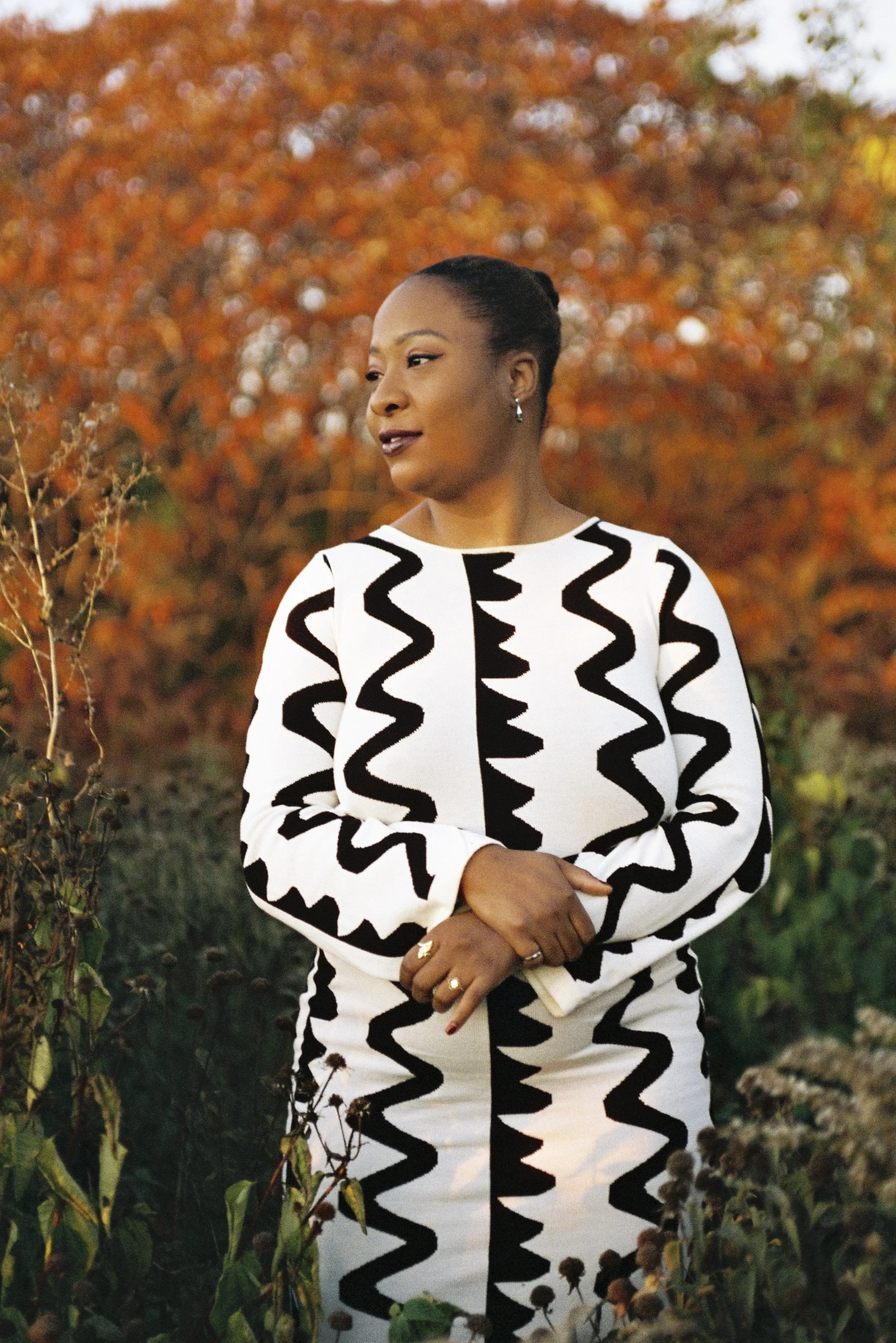 A woman with dark hair styled in a bun, wearing a black and white zigzag patterned dress, standing outdoors in front of autumn-colored foliage, with her hands crossed in front of her.