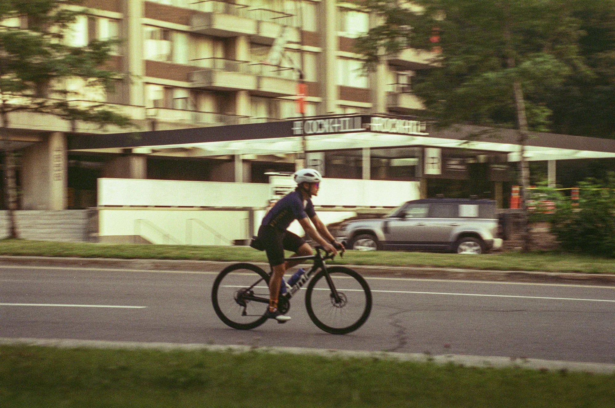 A person riding a bicycle wearing a helmet, riding along a city street with a building and cars in the background.