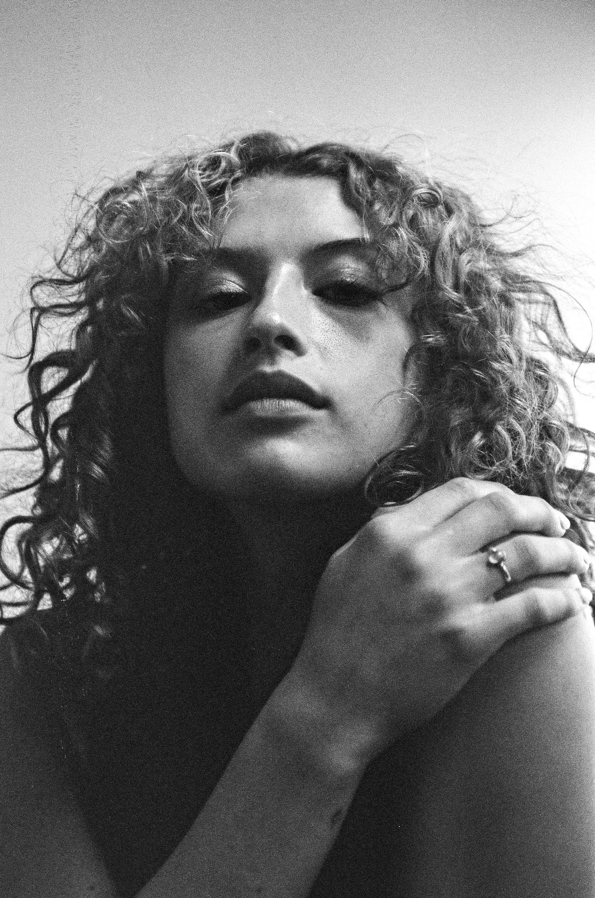 Close-up black and white photo of a woman's face with curly hair, lips slightly parted, and hand resting near chin, wearing a ring.