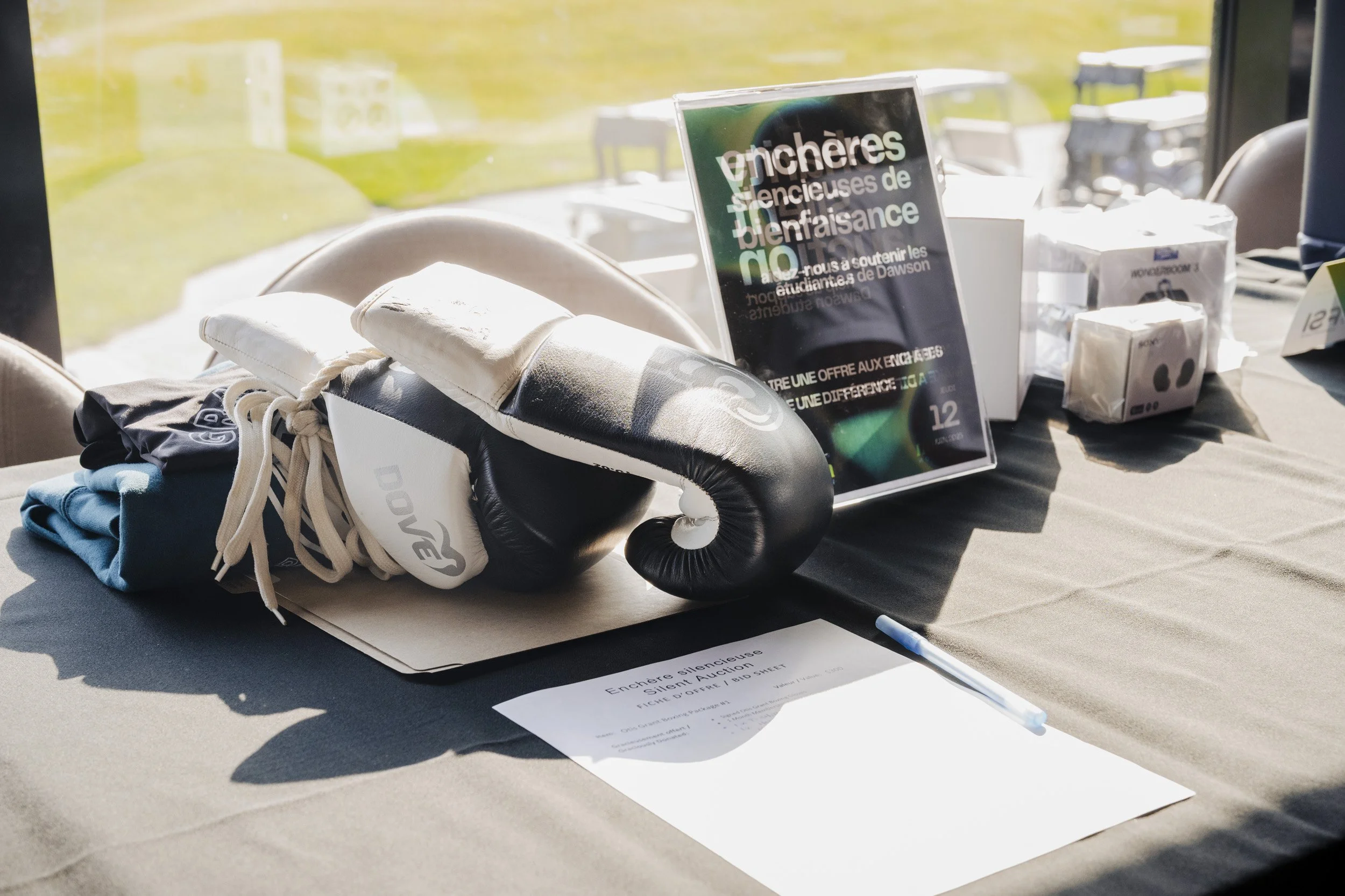 A table displaying boxing gloves, a face mask, folded clothing, and a sign in French about a silent auction for charity, with sunlight casting shadows on the table.