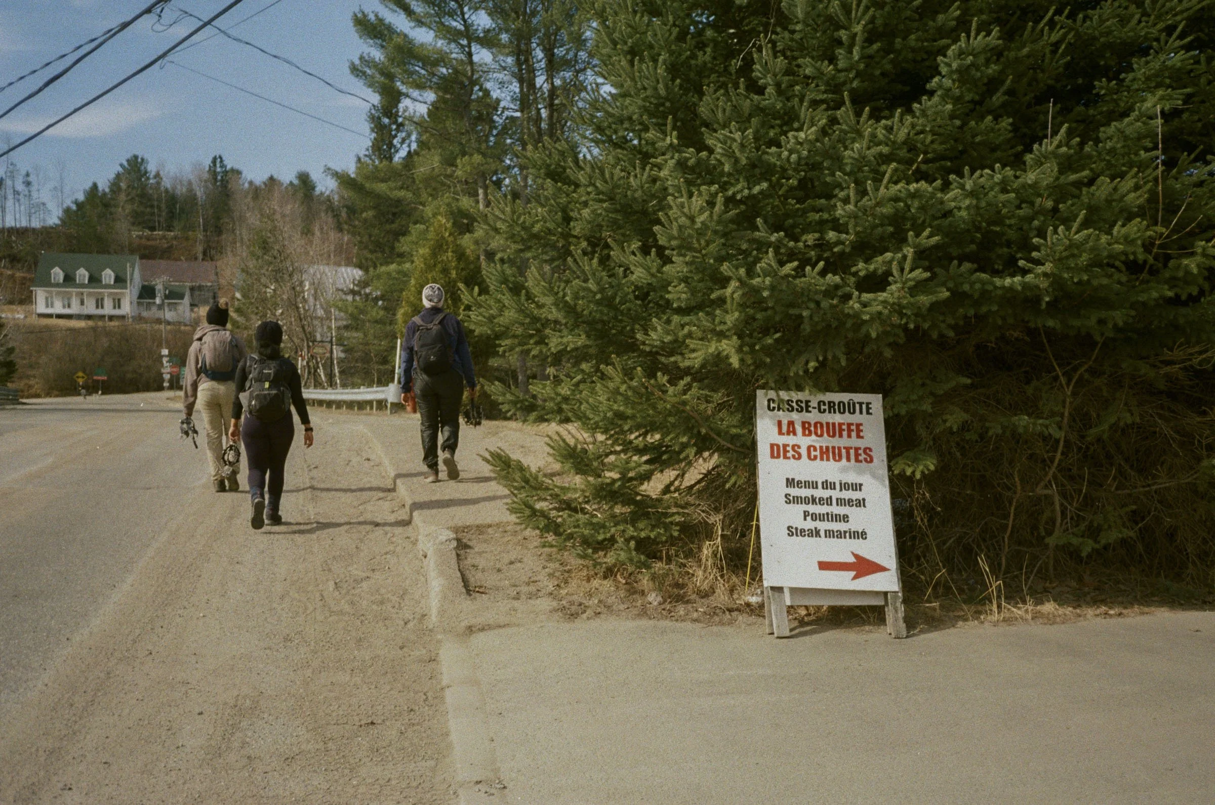 Three hikers walking along a sidewalk in a small town, with a large evergreen tree nearby and a sign listing meat options in French and English, including smoked meat, poutine, and steak mariné.