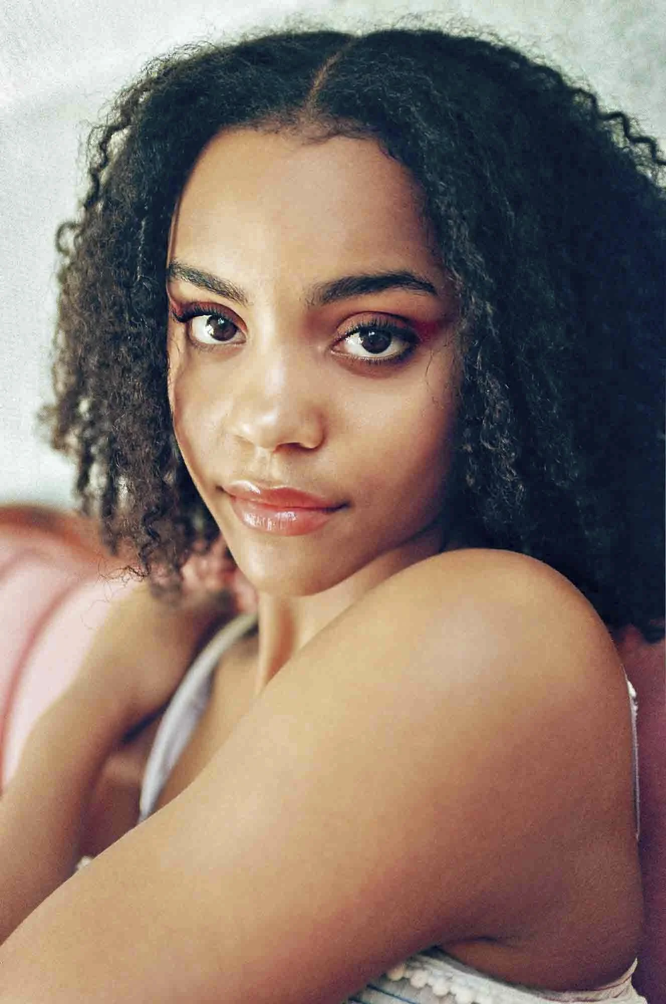 A young woman with curly dark hair and brown eyes, smiling softly, sitting on a bed with her arm crossed over her shoulder, wearing a sleeveless top, in a close-up portrait.