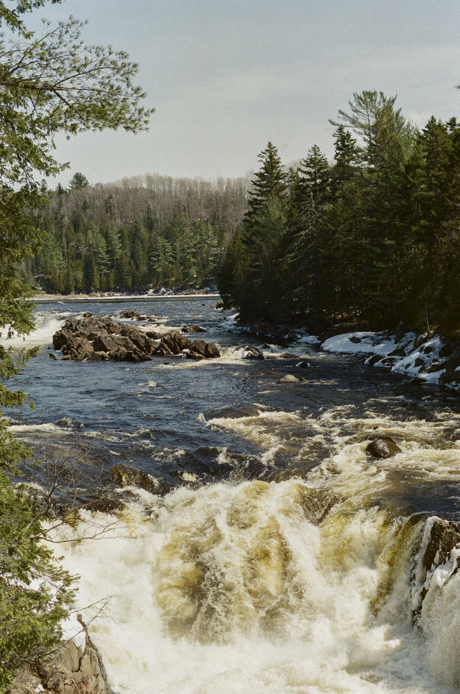 A river flowing through a forested area with trees on both sides and some snow on the ground.