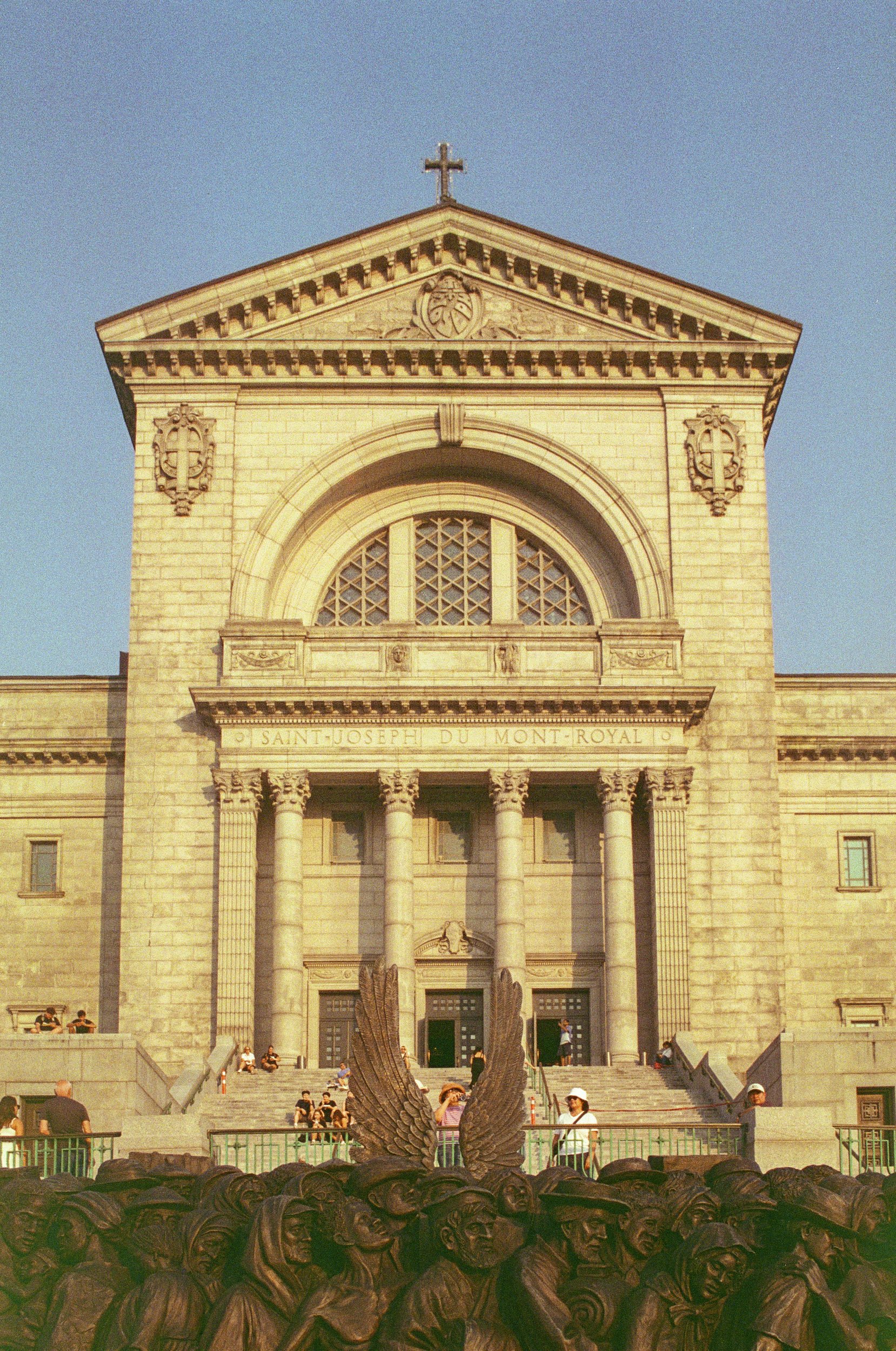 People gather in front of a large stone church with a triangular pediment, columns, and a cross on top. In the foreground, there is a metal sculpture of multiple human faces and a large eagle figure with outstretched wings.