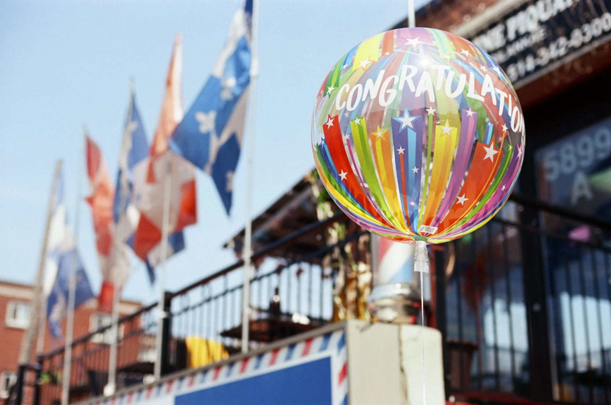 Colorful balloon with 'CONGRATULATIONS' written on it, hanging outdoors in front of a store with flags and a clear sky.