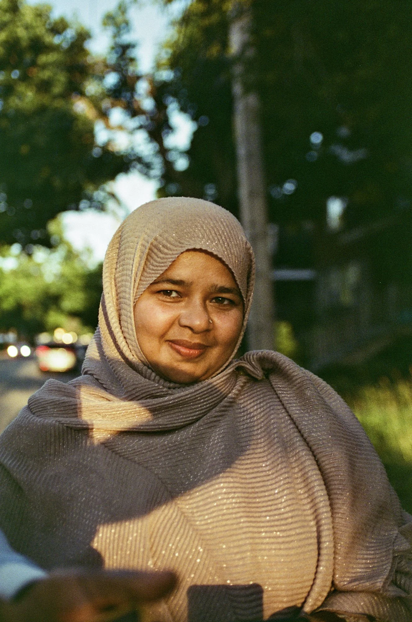 A woman wearing a beige hijab and a brown shawl is smiling outdoors during daytime, with blurred trees and cars in the background.