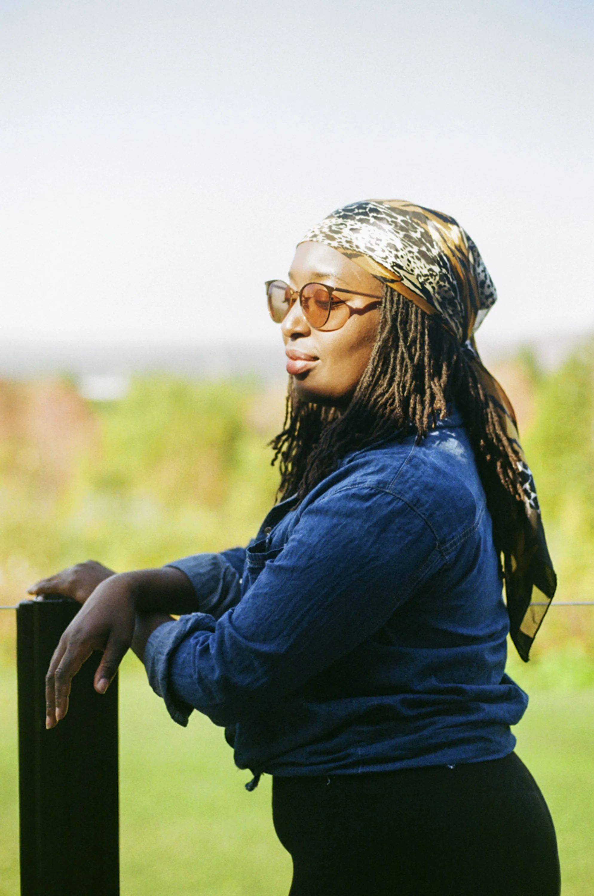 A woman with dreadlocks, wearing sunglasses, a patterned headscarf, and a denim jacket, standing outdoors near a black railing with greenery in the background.