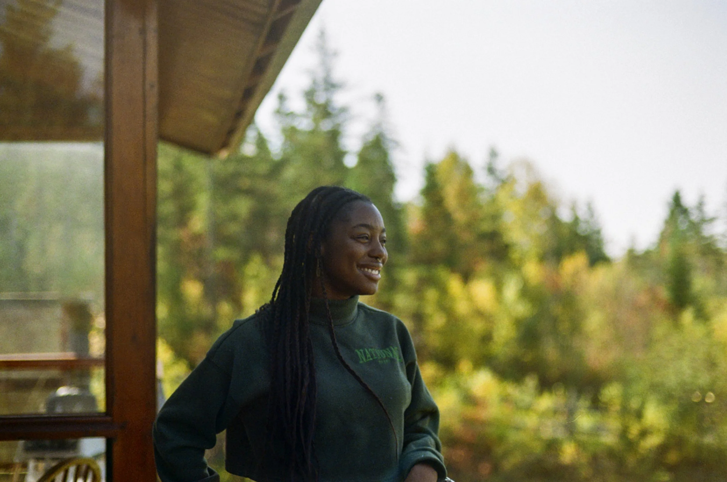 Smiling woman with braids standing outdoors near a wooden structure, with trees and sky in the background.