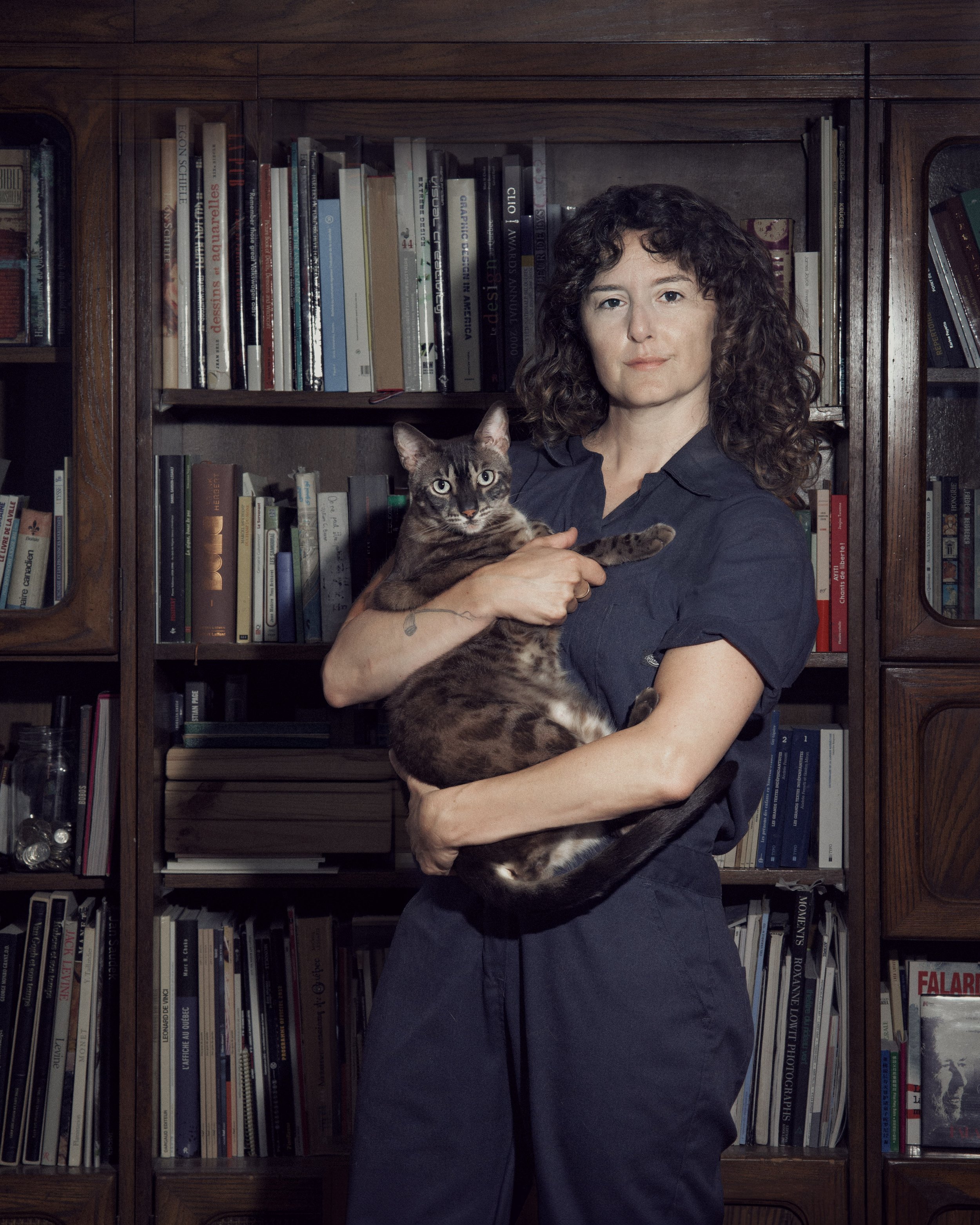 A woman with curly brown hair holding a tabby cat in front of a wooden bookshelf filled with books.