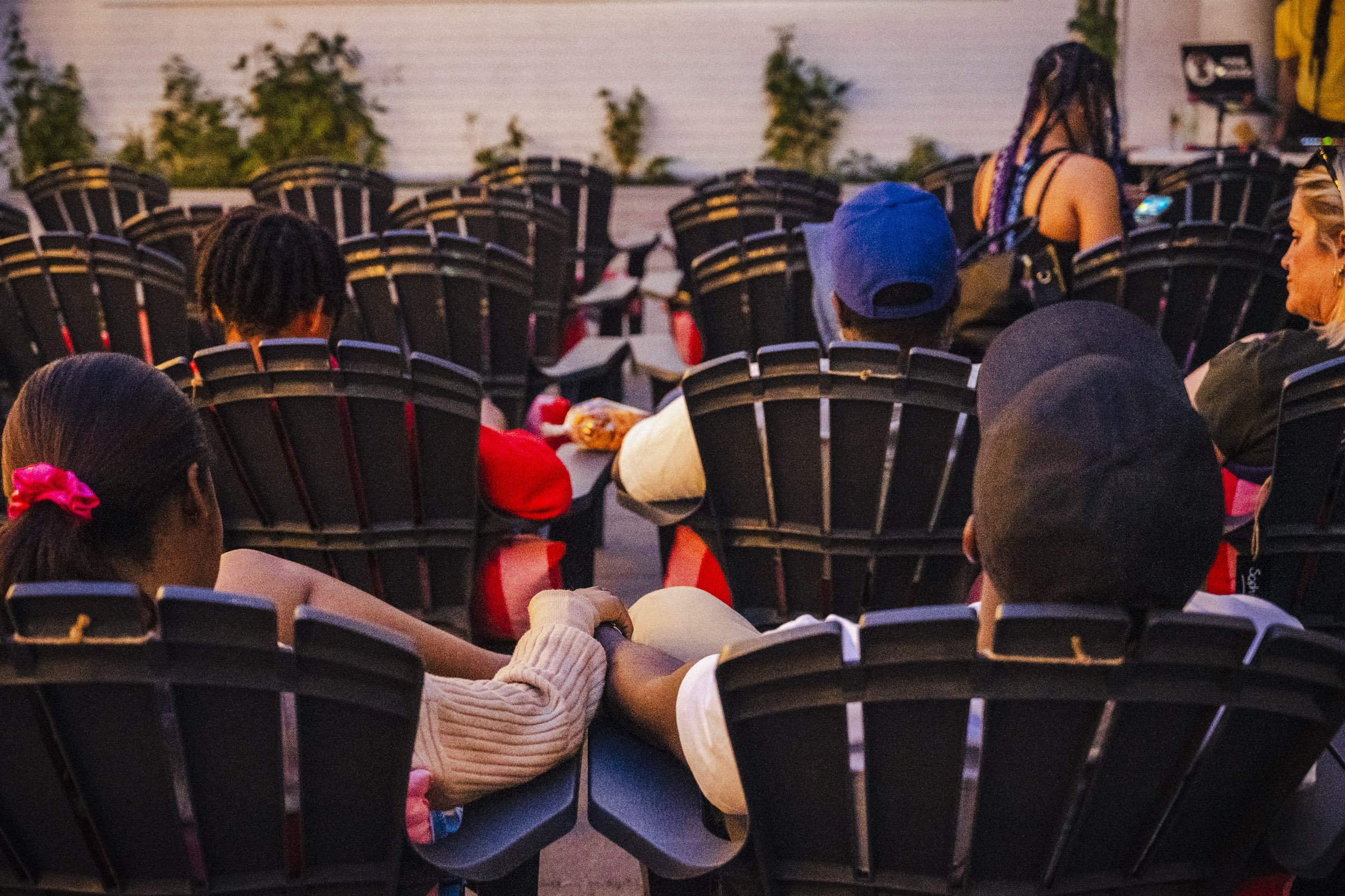 Audience members seated in black chairs at an event, some using phones, in an outdoor setting during dusk.