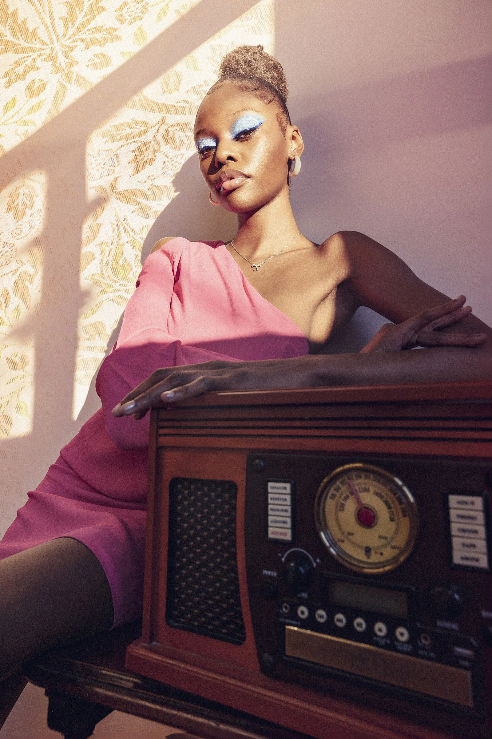 A woman with fair skin and styled hair wearing blue eyeshadow and pink lipstick, dressed in a pink dress and jewelry, leaning on a vintage radio and a piano, with patterned wallpaper and sunlight creating shadows in the background.