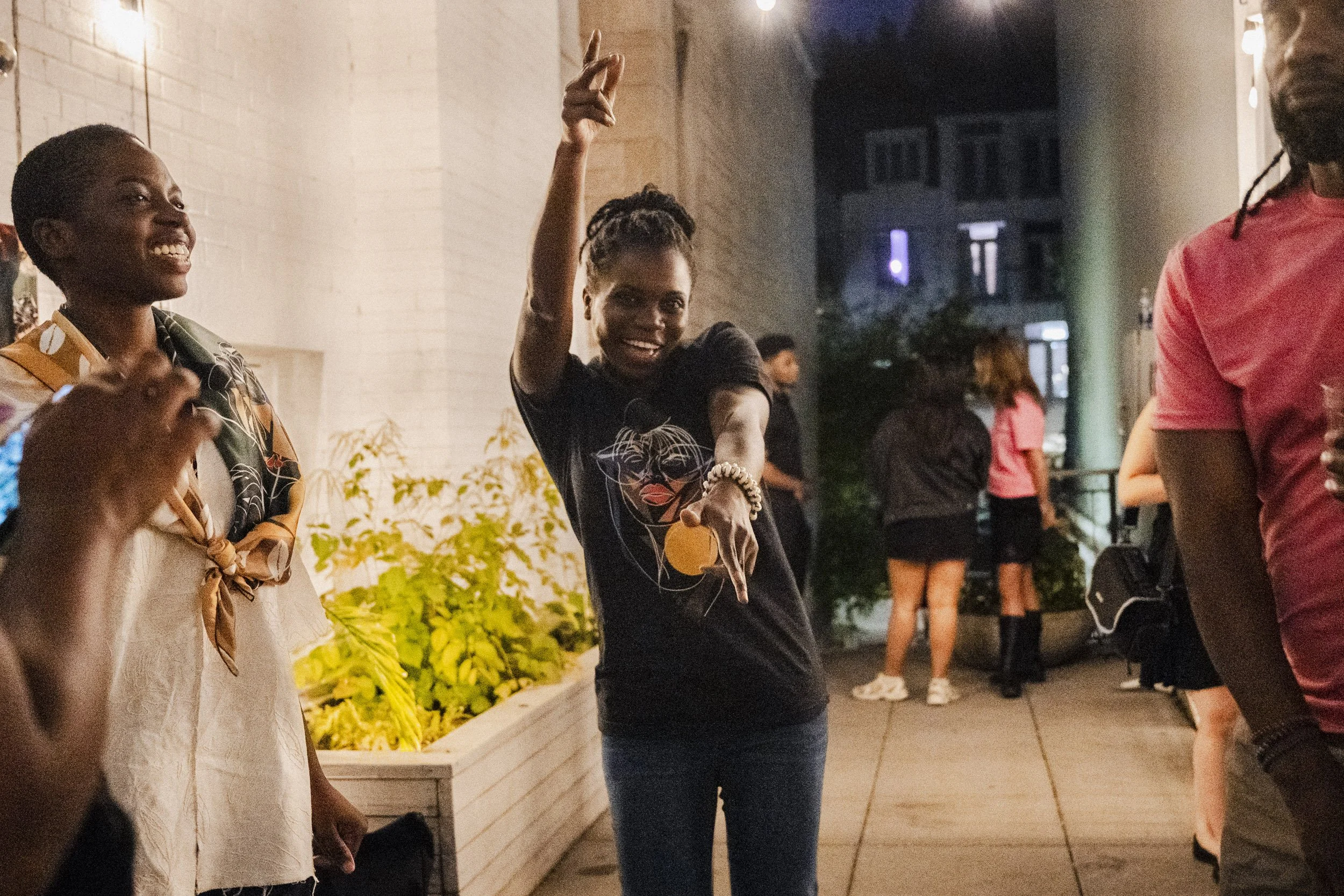 A woman in black shirt with graphic design pointing with an orange object in front of her, smiling, standing outdoors at night among people at a social gathering.