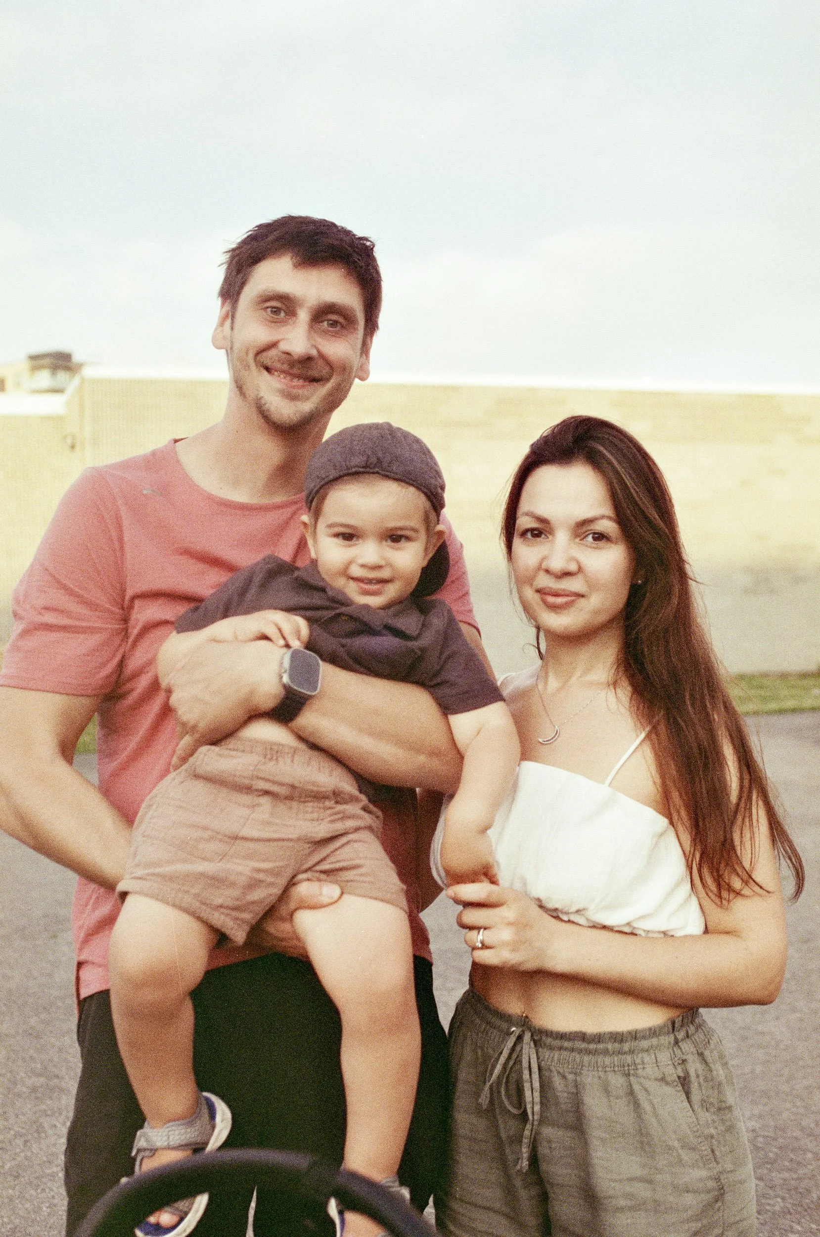 A family of three posing outdoors on a cloudy day: a man, woman, and a young boy. The man is holding the boy, and they are standing next to the woman.