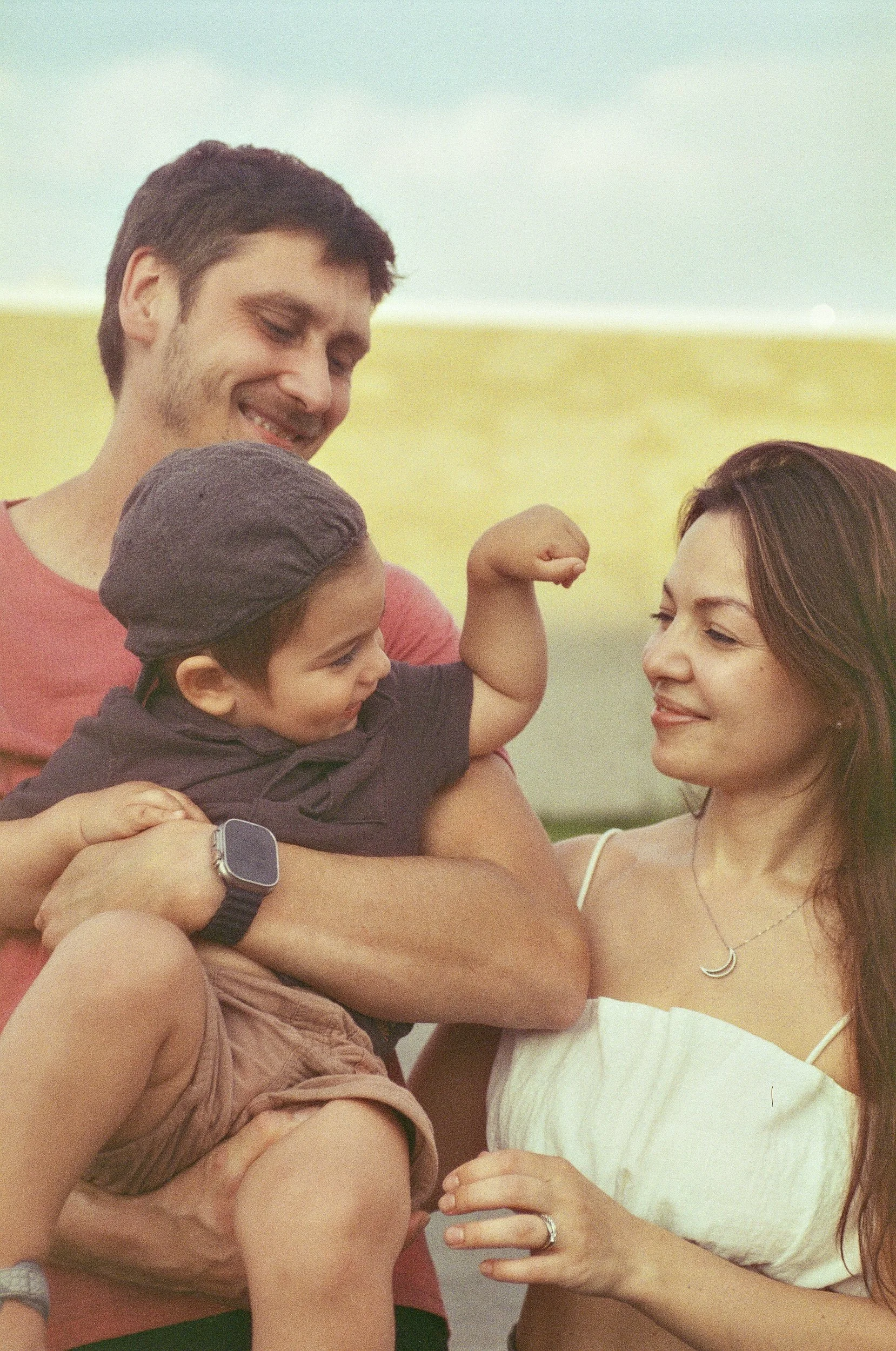 A smiling man and woman holding a young child outdoors during daytime, with open fields in the background.