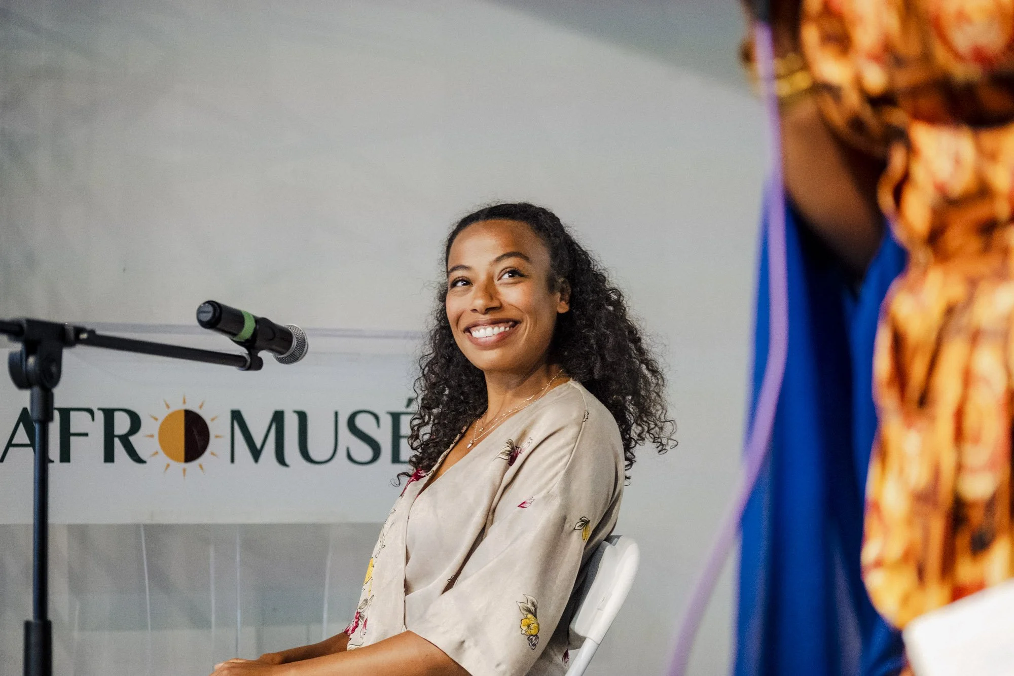 A woman with curly hair and a floral blazer smiling at a panel discussion. Behind her, part of a sign reads 'AFTER MUSE' and a microphone is positioned nearby.