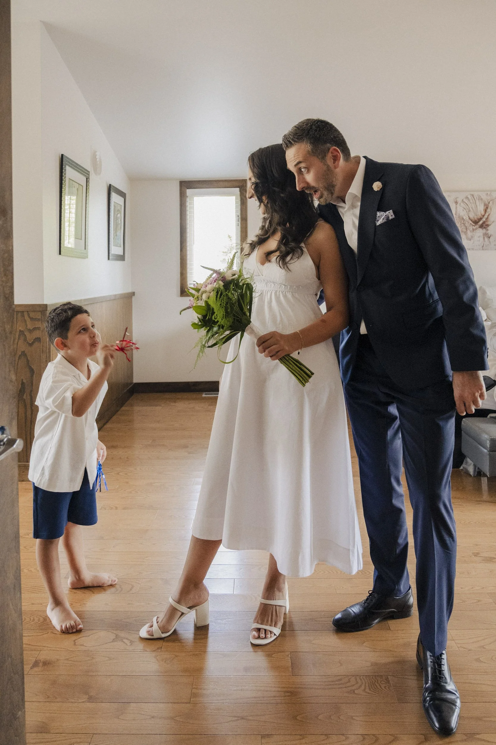 A woman in a white dress and beige high-heeled shoes holding a bouquet of flowers bends forward and talks to a man in a dark navy suit. The man is leaning forward, smiling, and wearing black shoes. A young boy with short dark hair, barefoot, wearing 