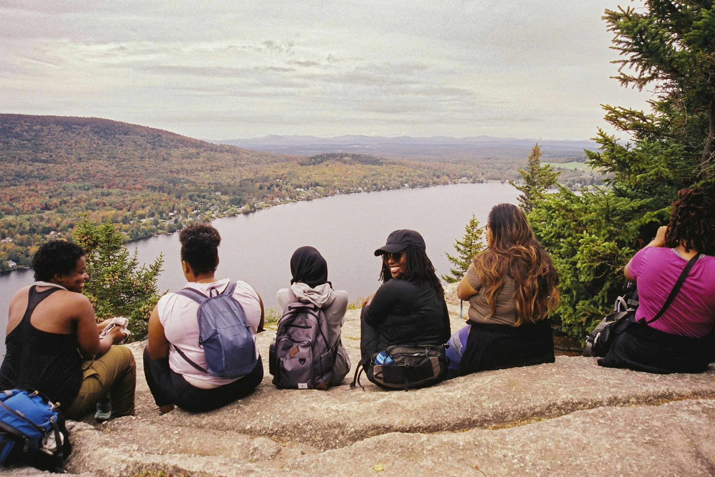 A group of diverse people sitting on a rock ledge overlooking a river and colorful autumn trees on hills, with cloudy sky above.