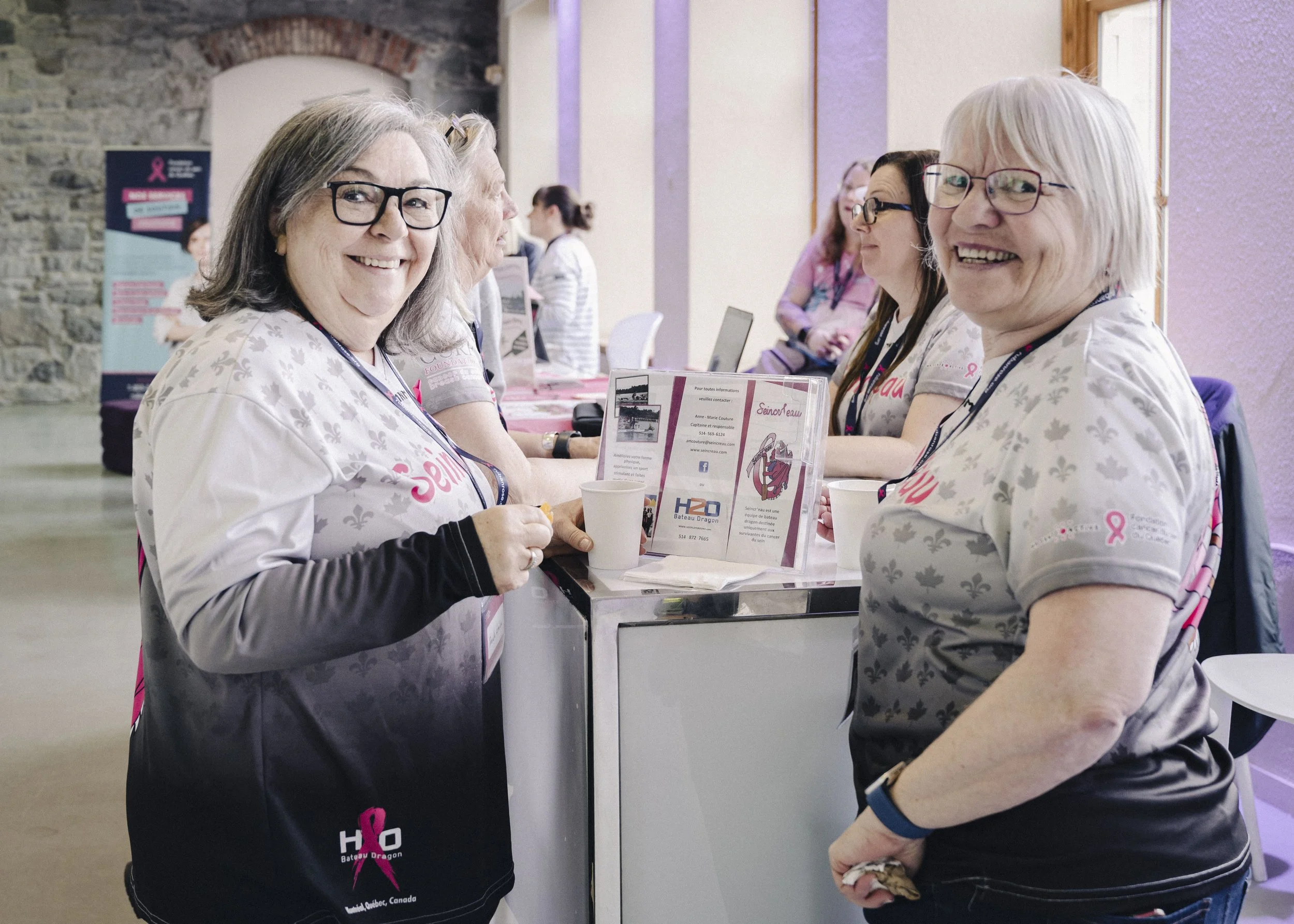 Smiling women at a table during a breast cancer awareness event, with informational brochures and pink ribbons, in a venue with stone and purple walls.