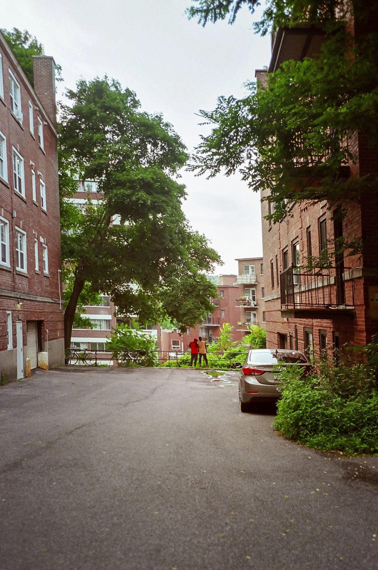 An outdoor residential courtyard surrounded by brick apartment buildings, with green trees, parked cars, and two people standing near a railing.