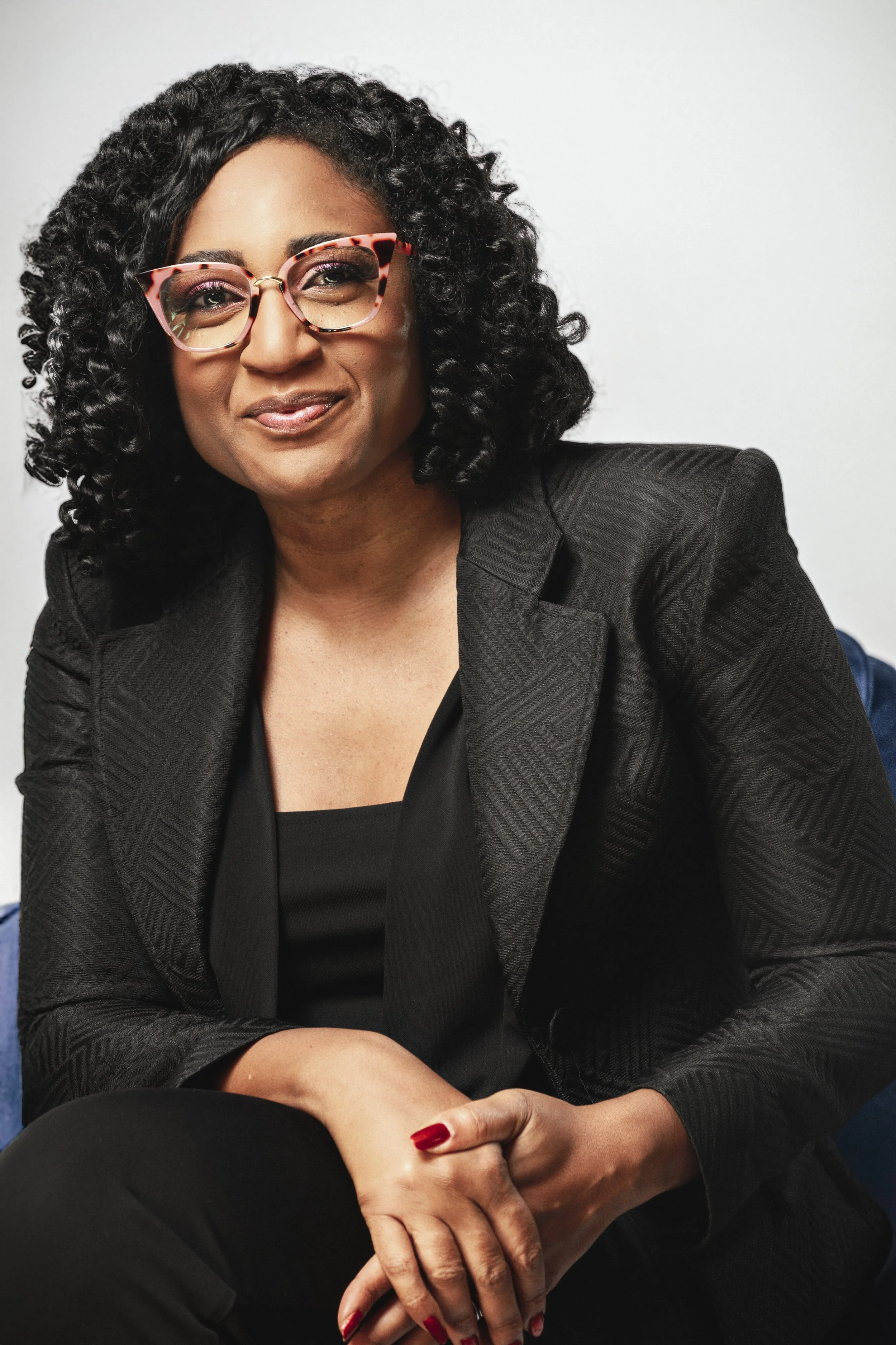 A woman with curly black hair, wearing pink tortoiseshell glasses, a black blazer, and red nail polish, sitting with her hands folded in her lap against a plain white background.