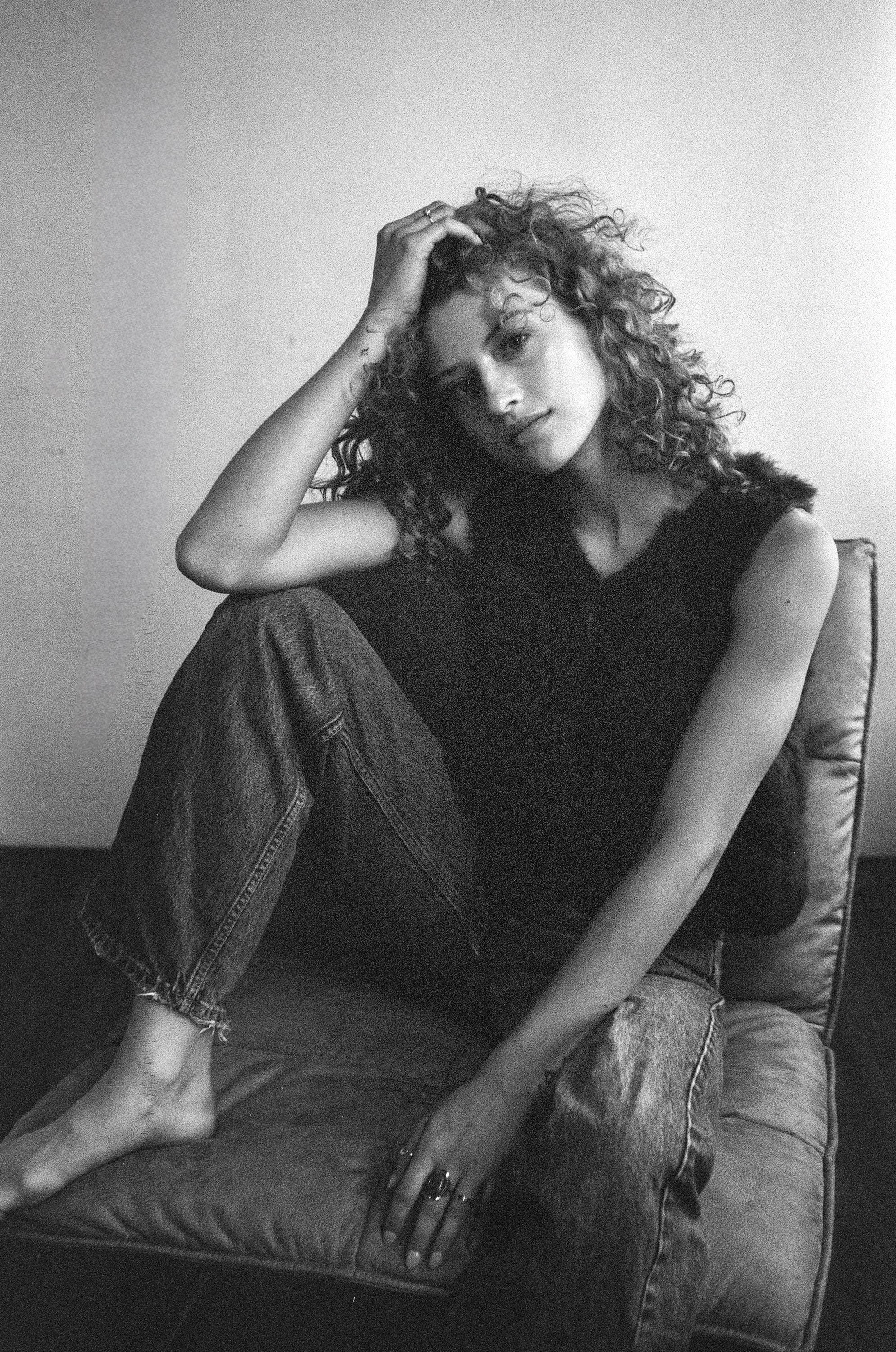 Black and white photo of a woman with curly hair sitting on a couch, resting her head on her hand, wearing a sleeveless top and ripped jeans.