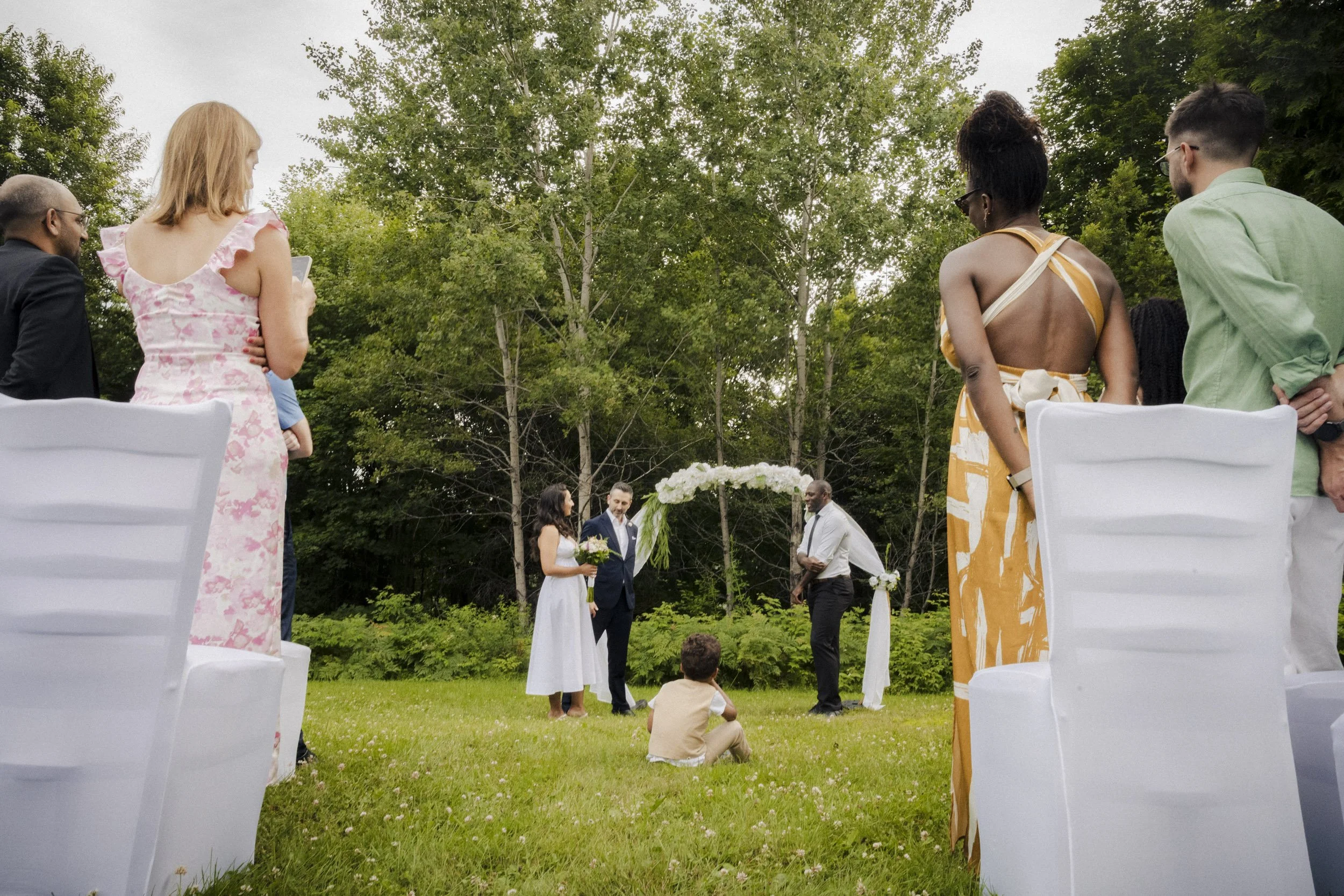 A group of people standing outdoors at a wedding ceremony, with the bride and groom in the center under a floral arch, surrounded by guests seated on white chairs on a grassy area with trees in the background.