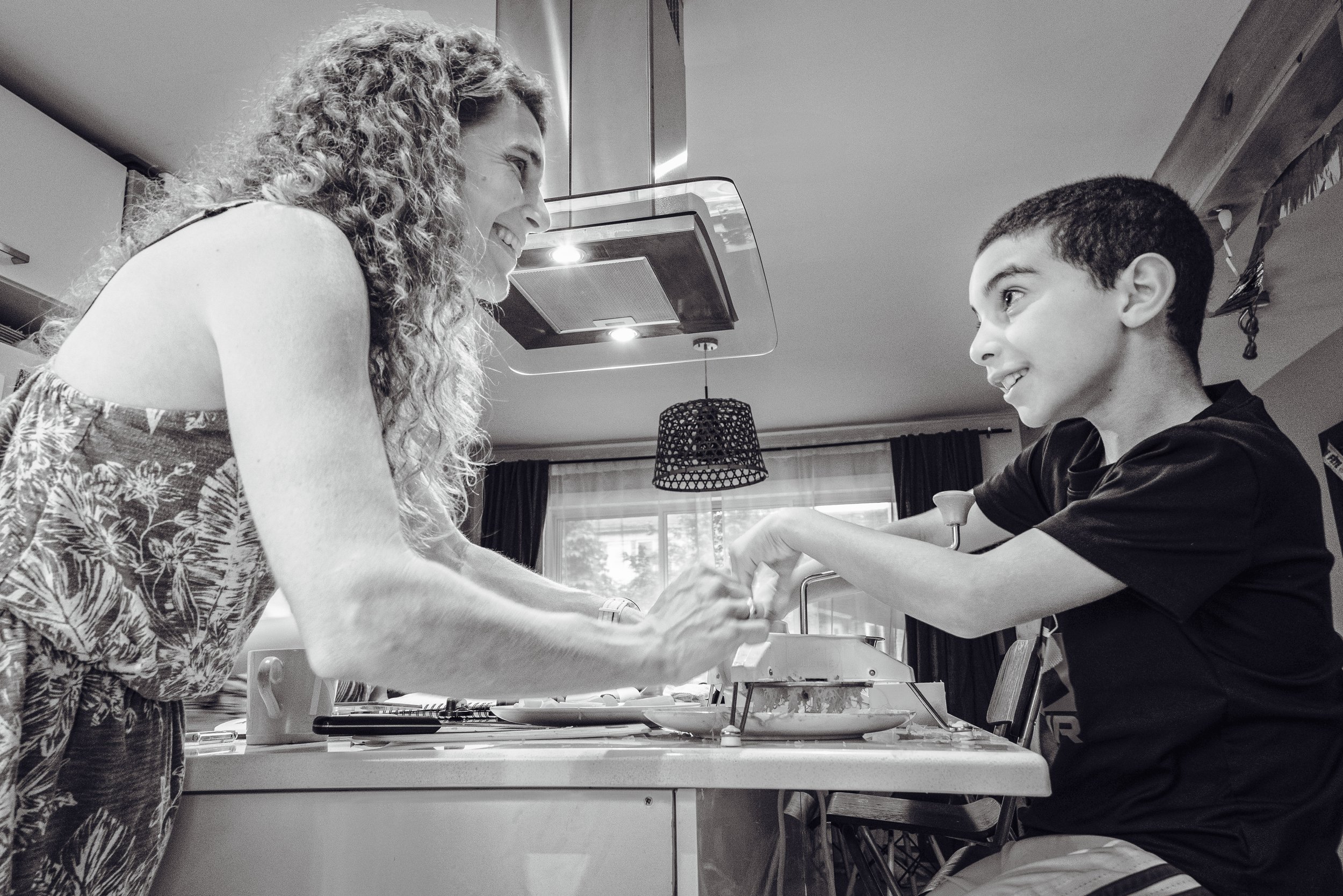 A woman and a young boy are looking at each other and smiling, holding hands across a table in a bright room, with kitchen elements visible in the background.