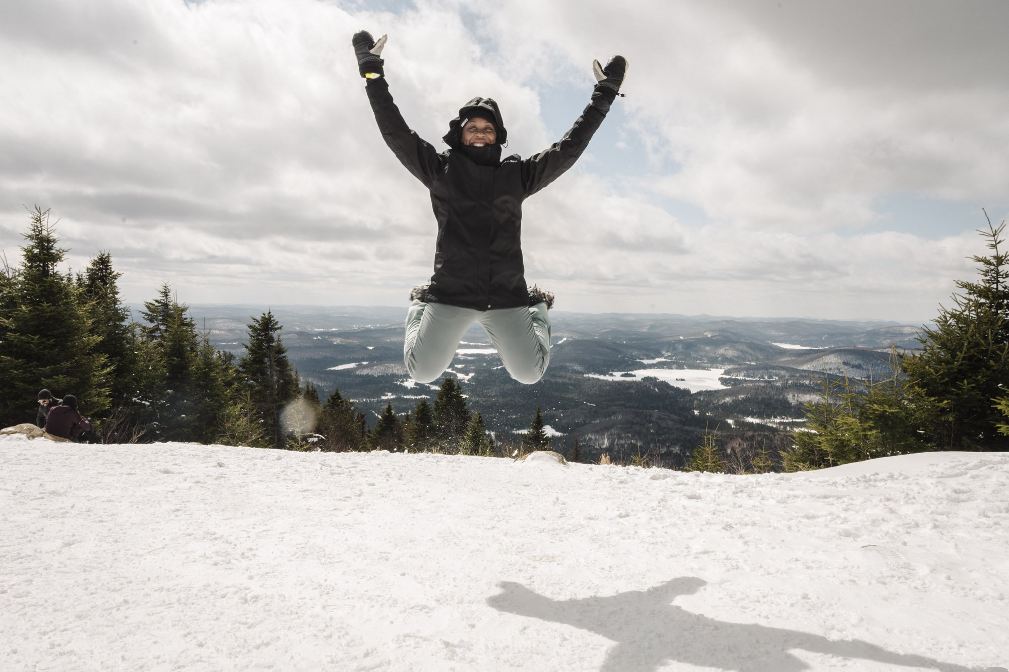 Person jumping in the air on a snowy mountain with a scenic view of forested hills and clouds in the sky.