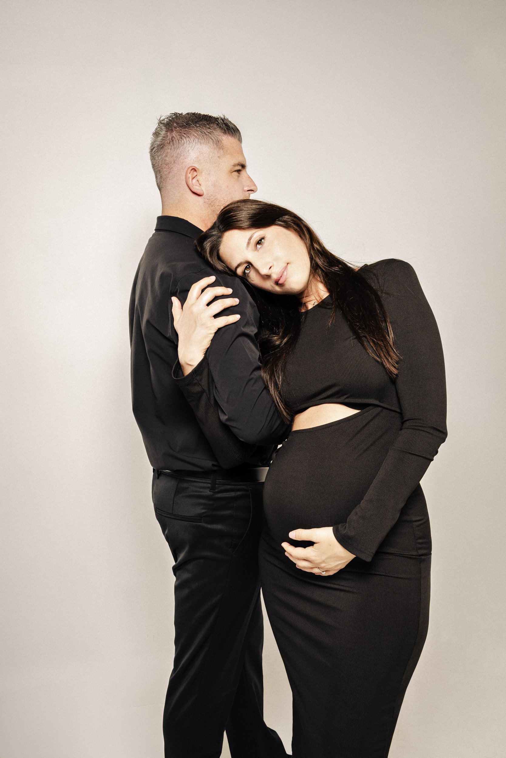 A pregnant woman with dark hair resting her head on a man's chest, who has short light hair, both dressed in black, against a plain beige background.