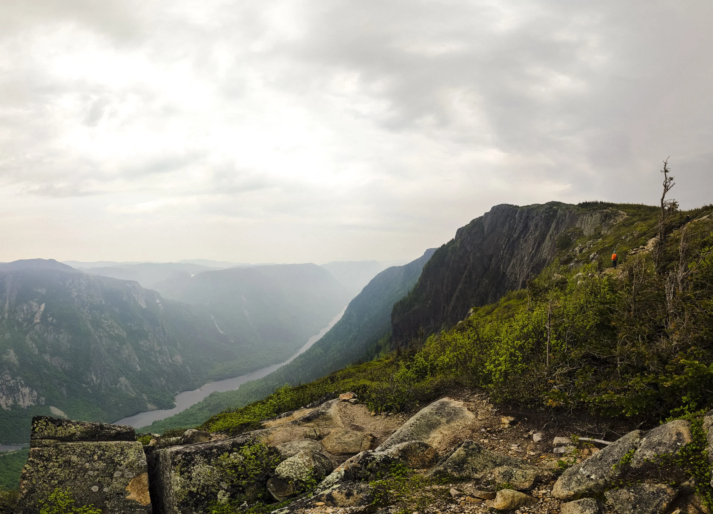 A scenic view of a valley with a river running through it, surrounded by green mountains and a cloudy sky. There are hikers walking along a trail on the right side of the image.