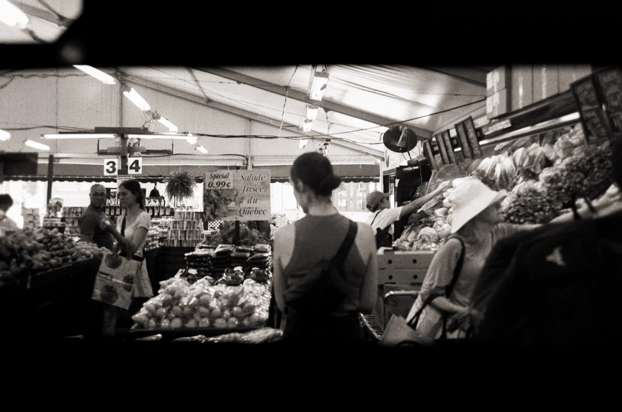 View of a marketplace stall with various fruits and vegetables, with people shopping and signs displaying prices, in a covered market space.