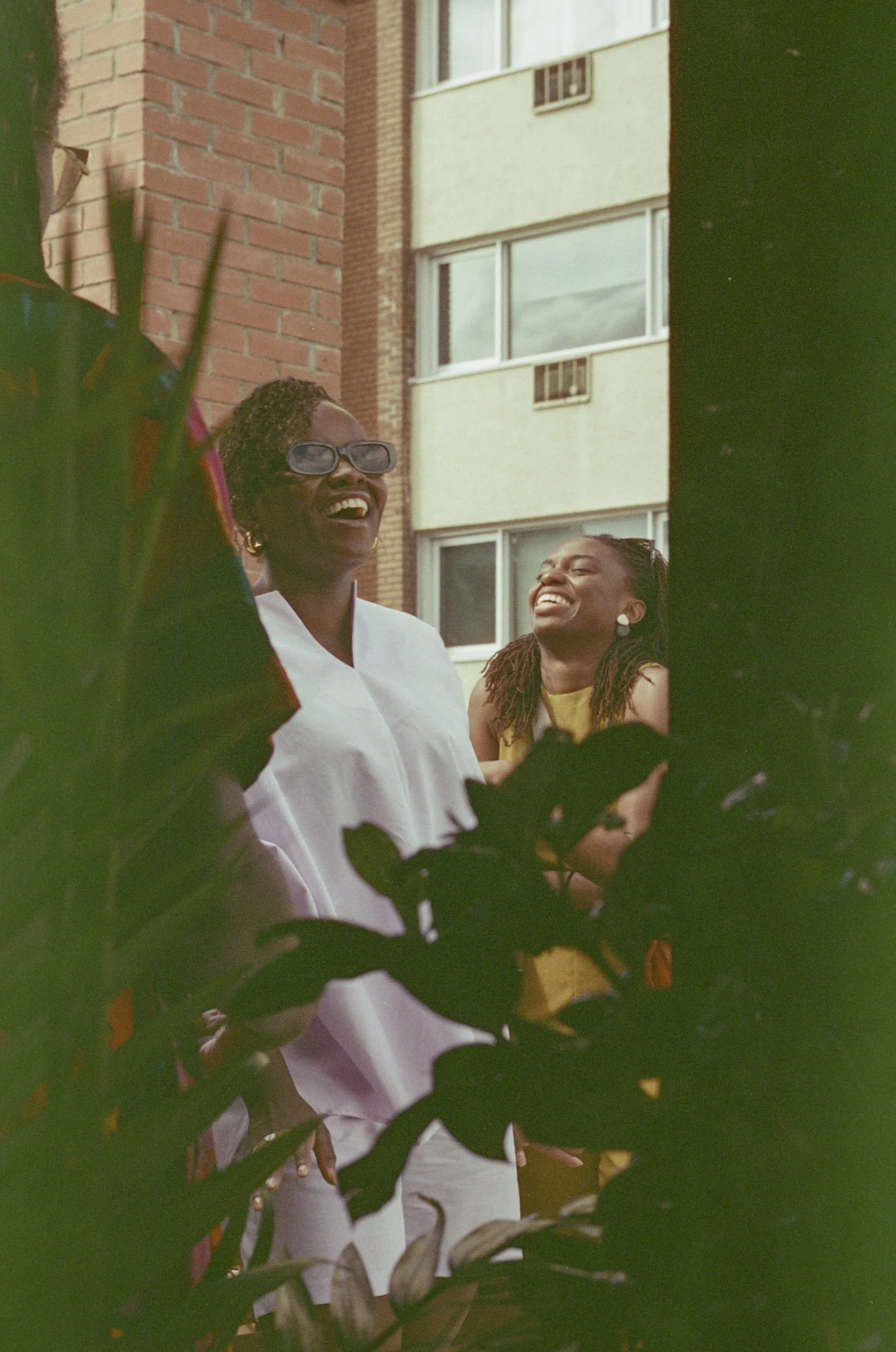 Two women laughing and smiling with a building in the background, framed by dark green leaves in the foreground.