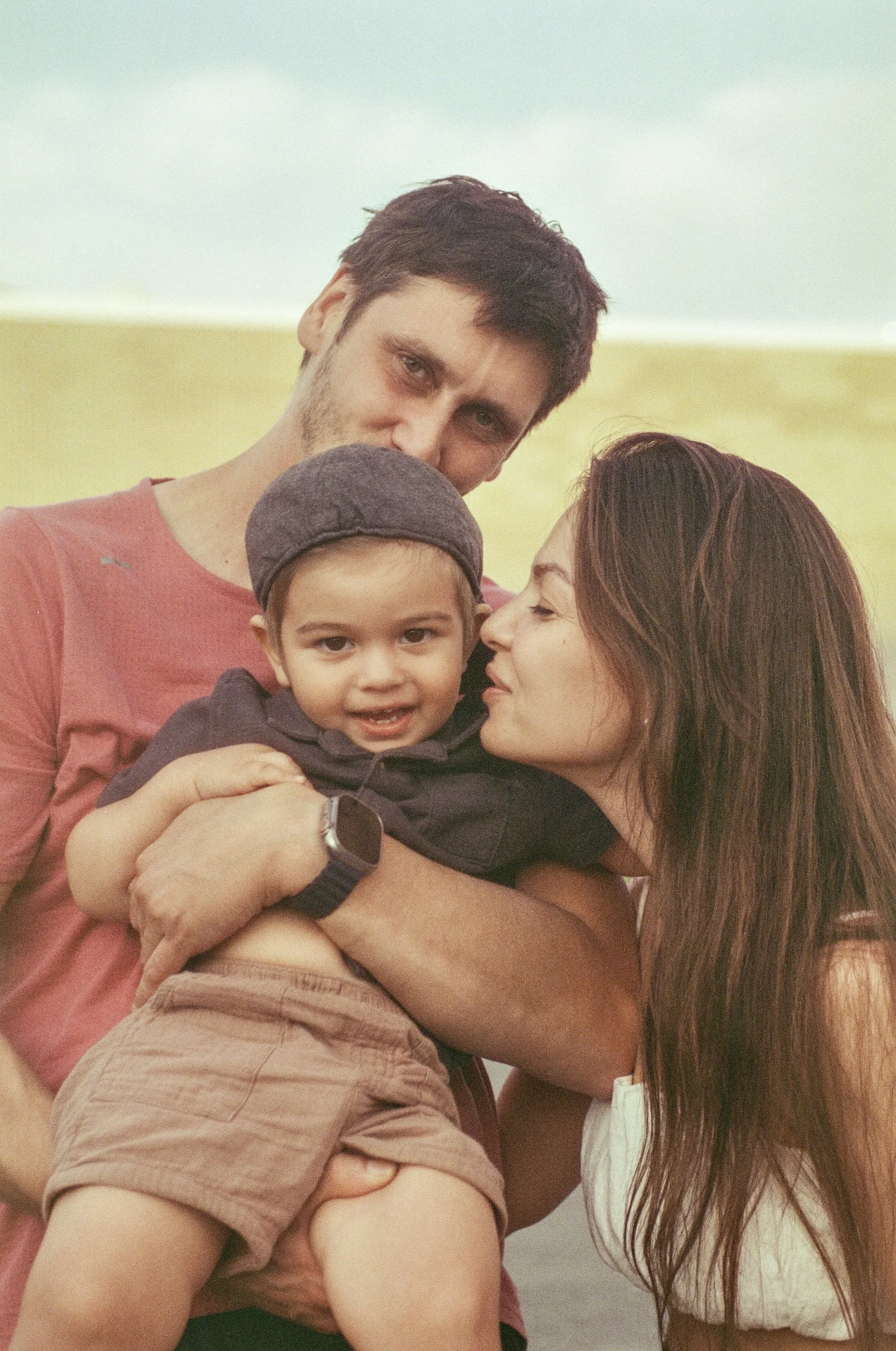 A family of three, including a man, a woman, and a young boy, close together at the beach with a pale sky and sand in the background.