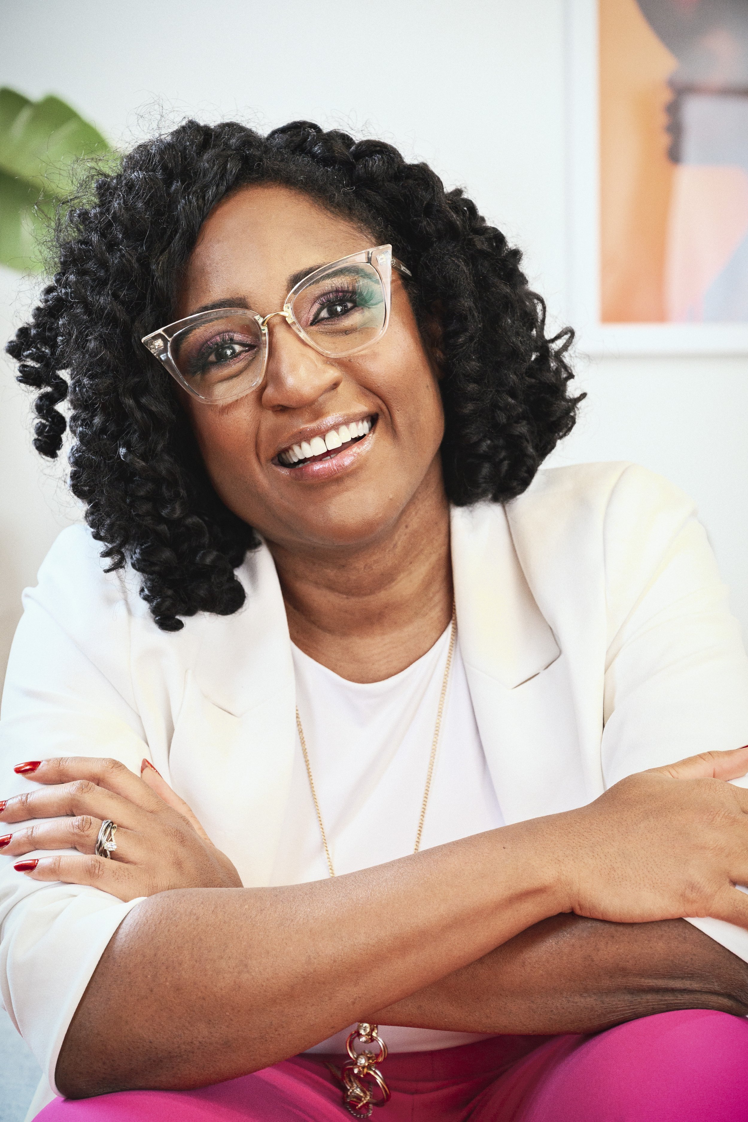 Close-up of a smiling woman with curly black hair, wearing clear glasses, a white blazer, and pink pants, sitting with arms crossed, in a bright, modern setting with a plant and abstract artwork in the background.