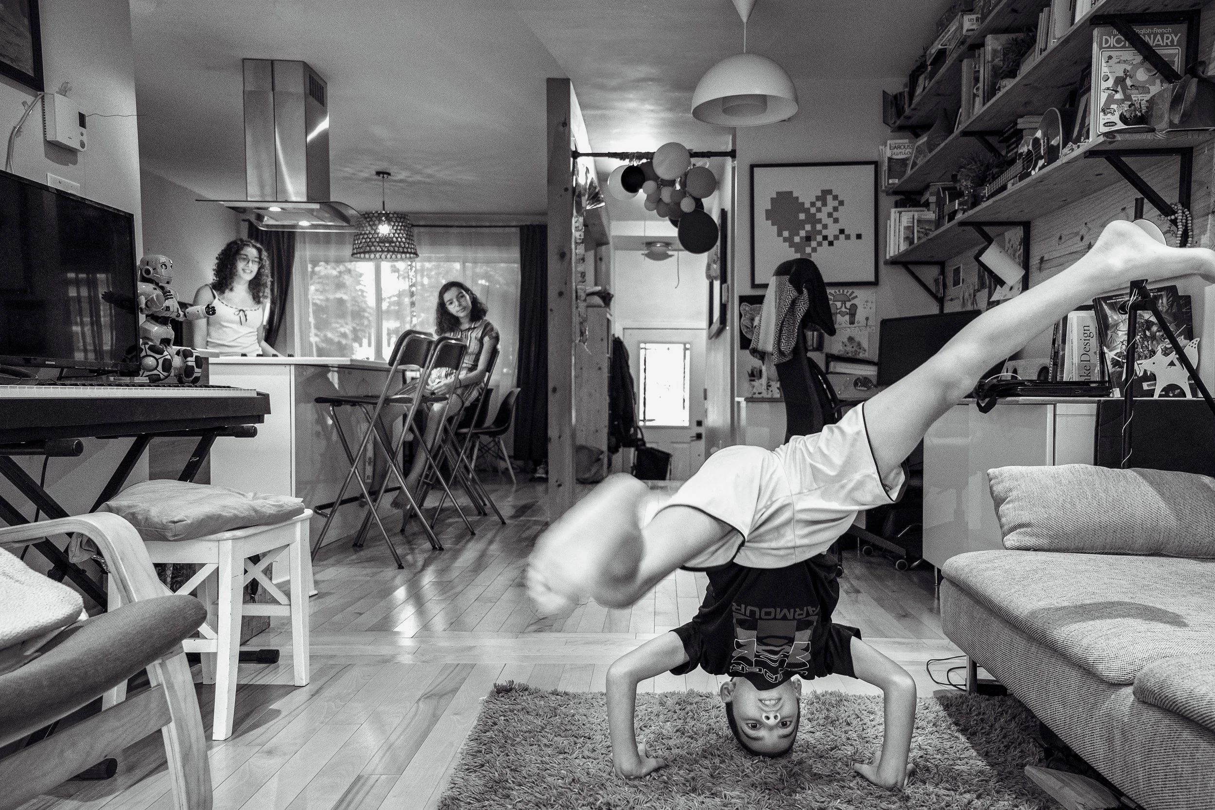 A child doing a handstand on a rug in a living room, with two women smiling and sitting at a table in the background, and various furniture, shelves, and artwork in the room.