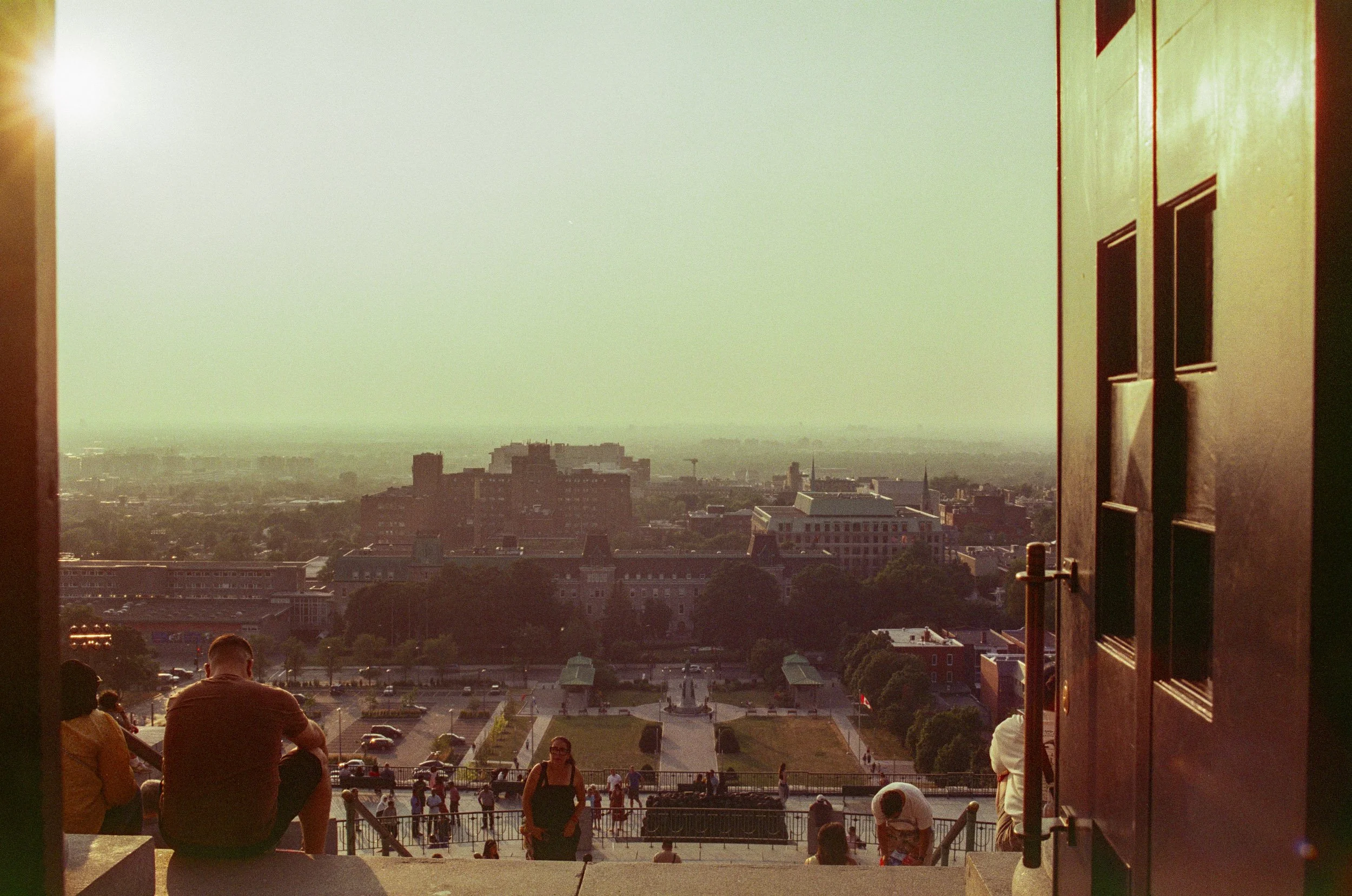 People sitting on stairs overlooking a cityscape and a large open square, with a hazy horizon and buildings in the distance, seen through a framed opening.