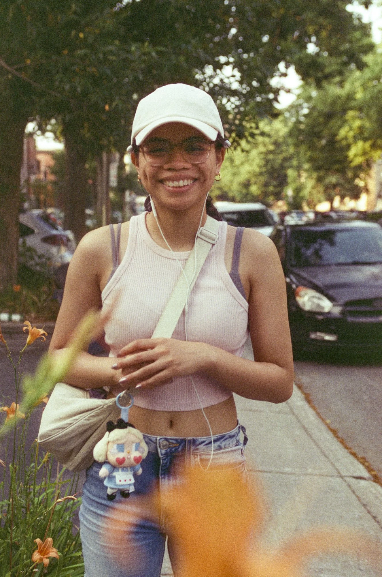 A smiling young woman wearing a beige baseball cap, glasses, a sleeveless striped pink top, and denim shorts, walking outdoors with greenery and parked cars in the background.