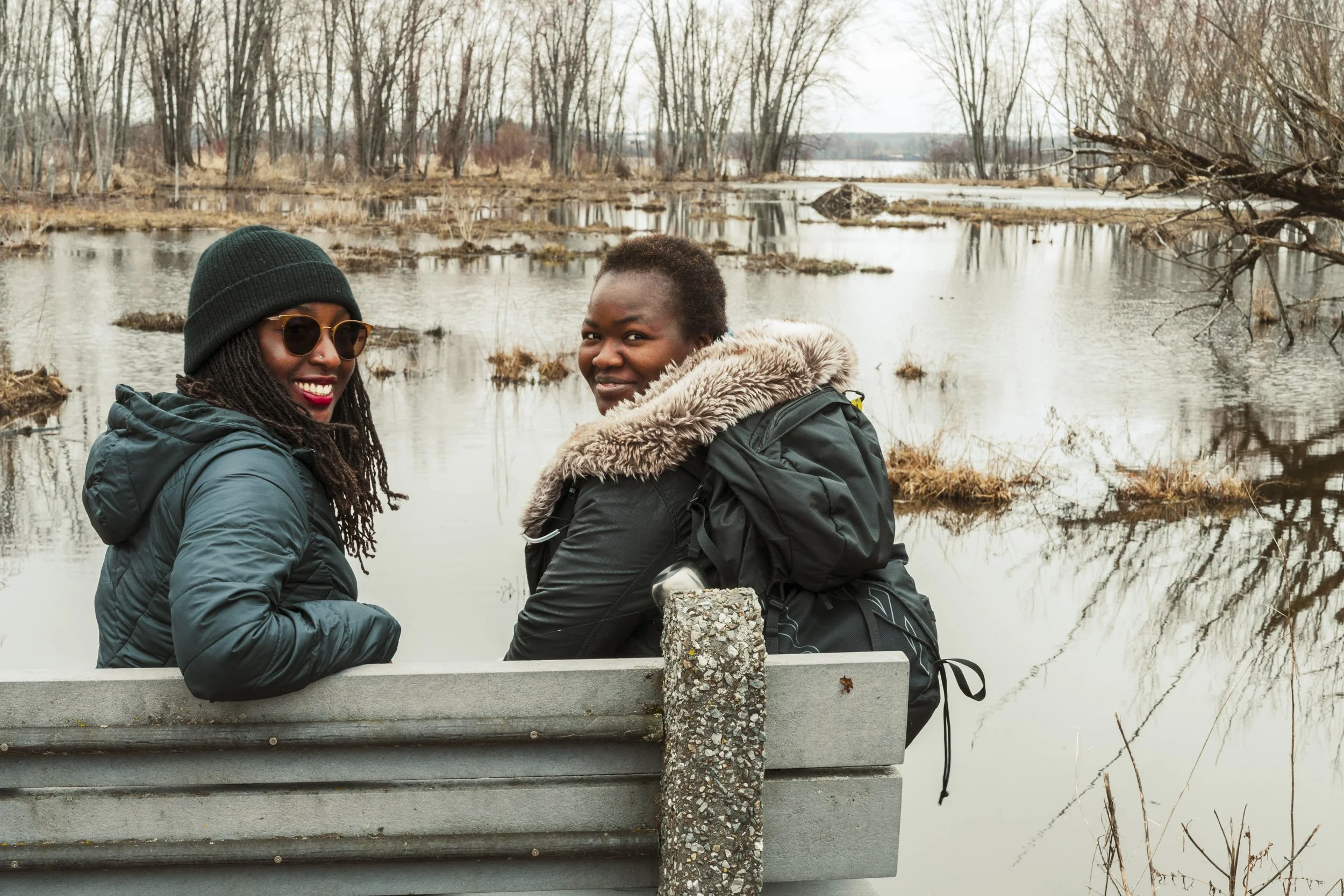Two women standing outdoors near a body of water, smiling at the camera. They are wearing winter jackets, with one in a black beanie and sunglasses, and the other with a fur-lined hood. The background features leafless trees and overcast sky.