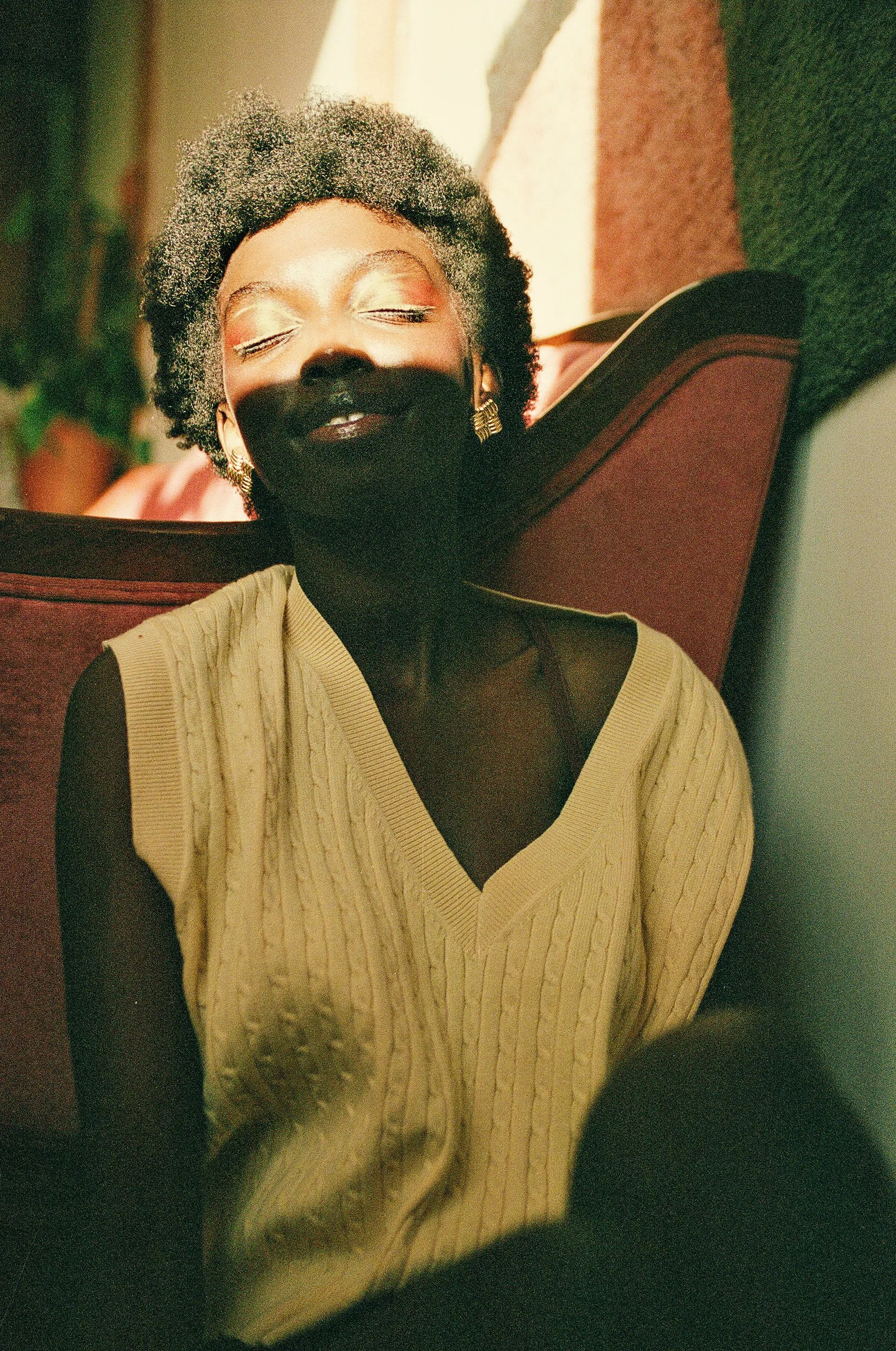 A woman with short curly gray hair and earrings, leaning back on a red cushioned chair with her eyes closed, wearing a beige sweater.
